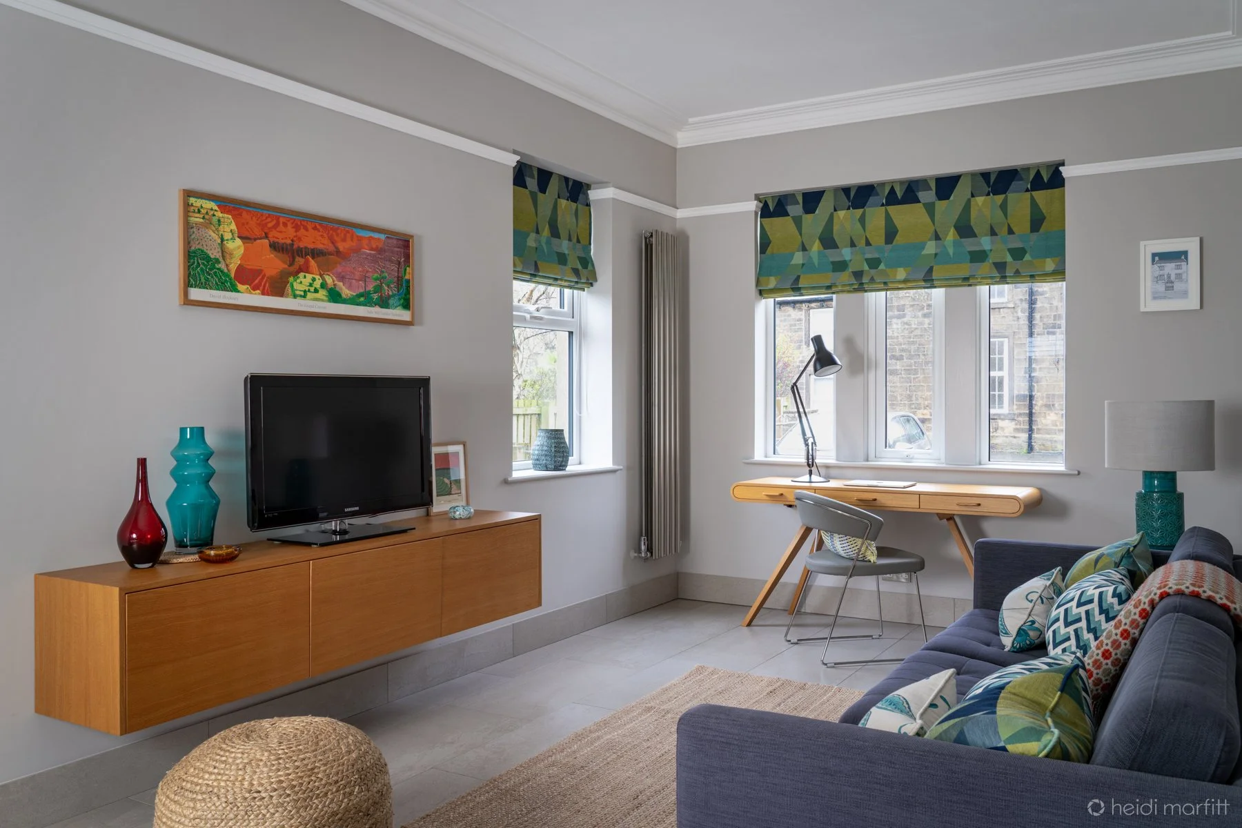 Living room with a gray wall, a wooden TV stand with vases and a TV, a wall art piece, a windowed corner with a desk and lamp, a gray sofa with colorful pillows, a lamp on side table, and a woven pouf.