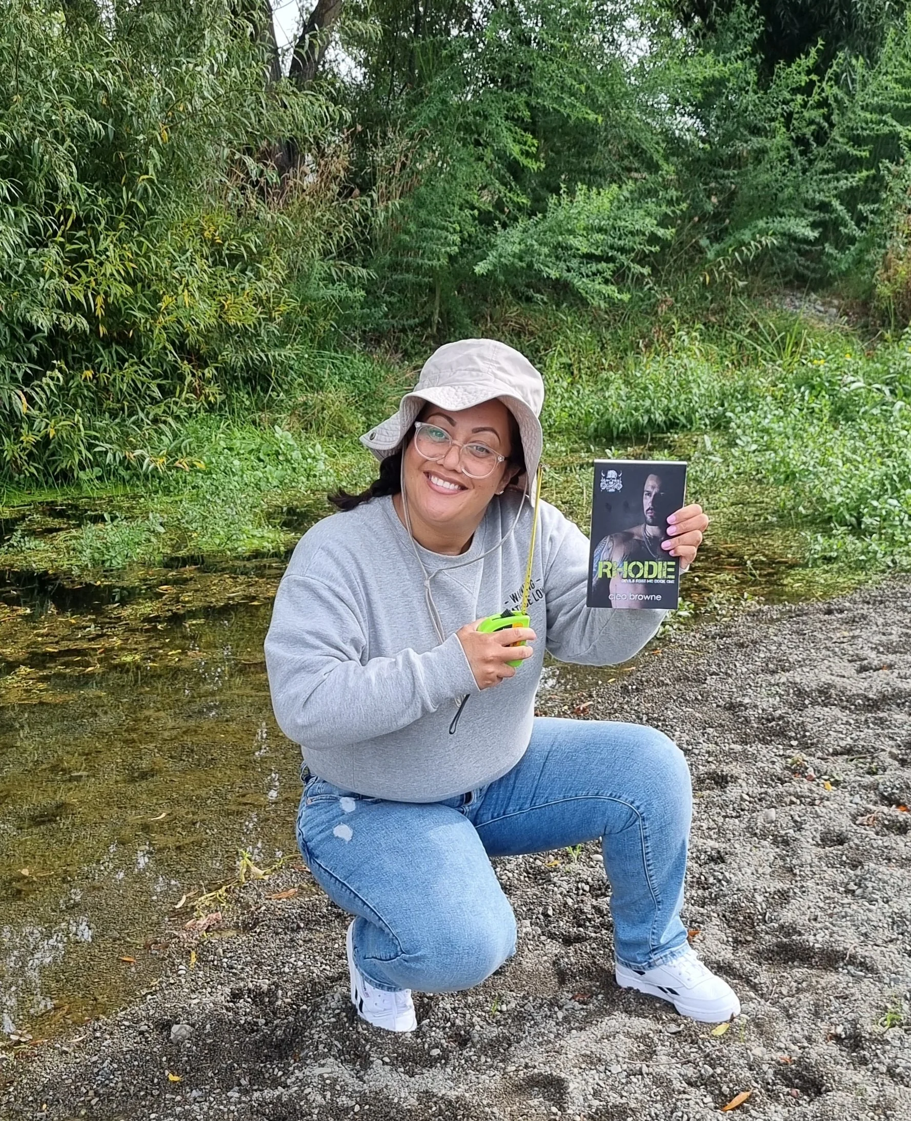 A woman kneeling on a gravelly riverbank, holding a small device in one hand and a promotional card featuring a man's face in the other, with lush green trees and shrubs in the background.