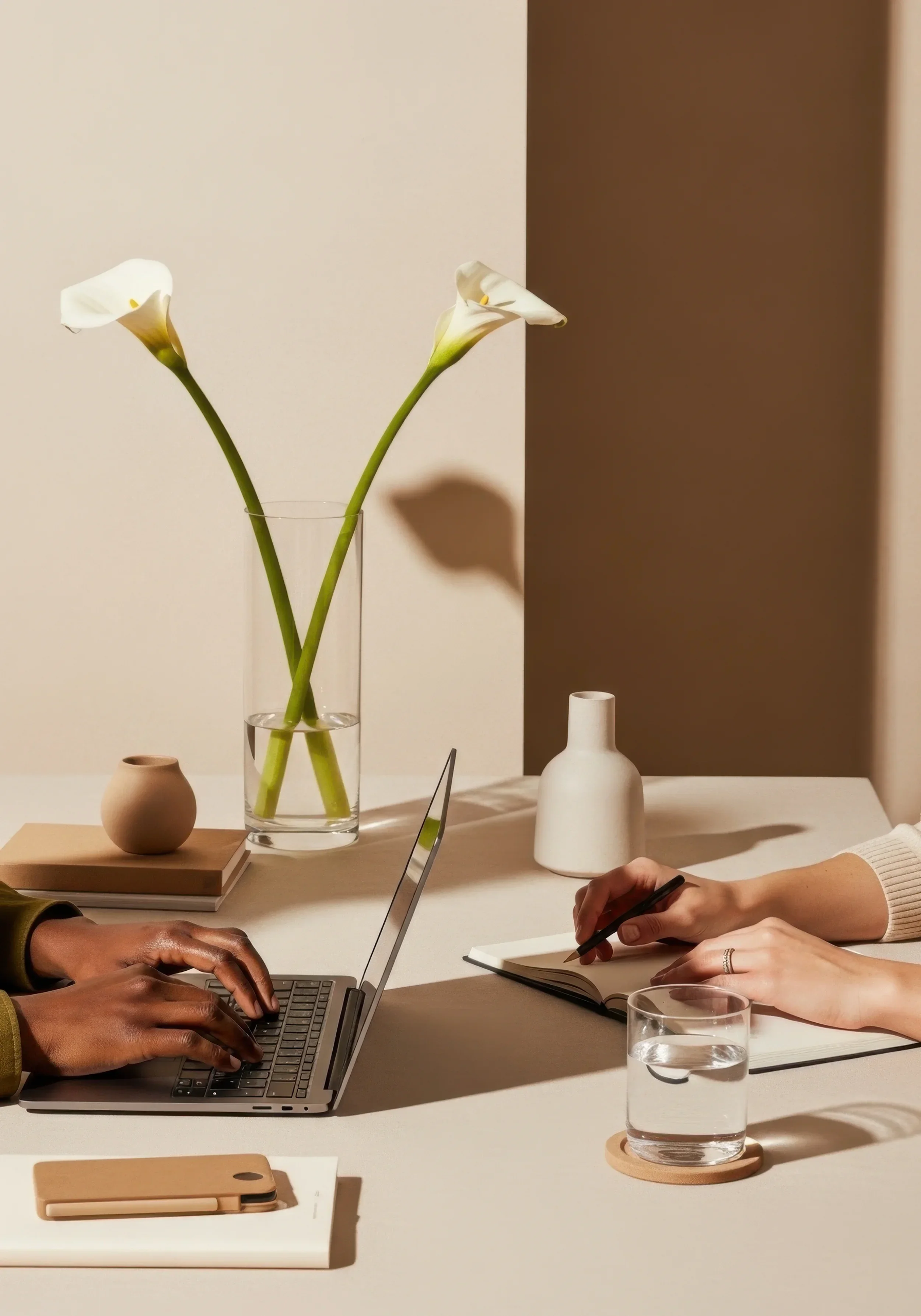 Cala lily in a glass vase next to two people working at a table with laptop, notebook, and a glass of water, and other items.