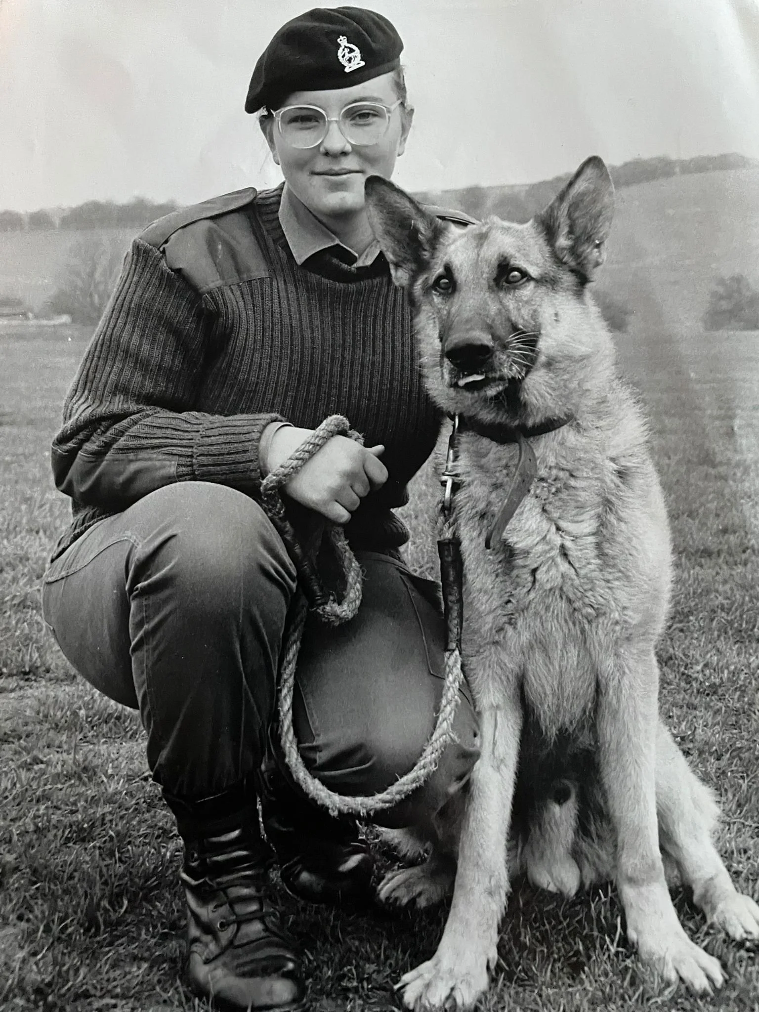 A woman in military attire with a beret kneeling outdoors with a German Shepherd dog on a leash.