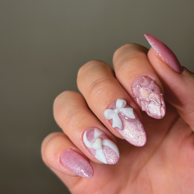 Close-up of a hand with pink, glittery manicured nails featuring white 3D bow decorations.
