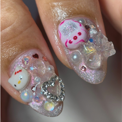Close-up of decorated fingernails with pink, white, and silver-themed nail art, featuring Hello Kitty and bear charms, rhinestones, and glitter.