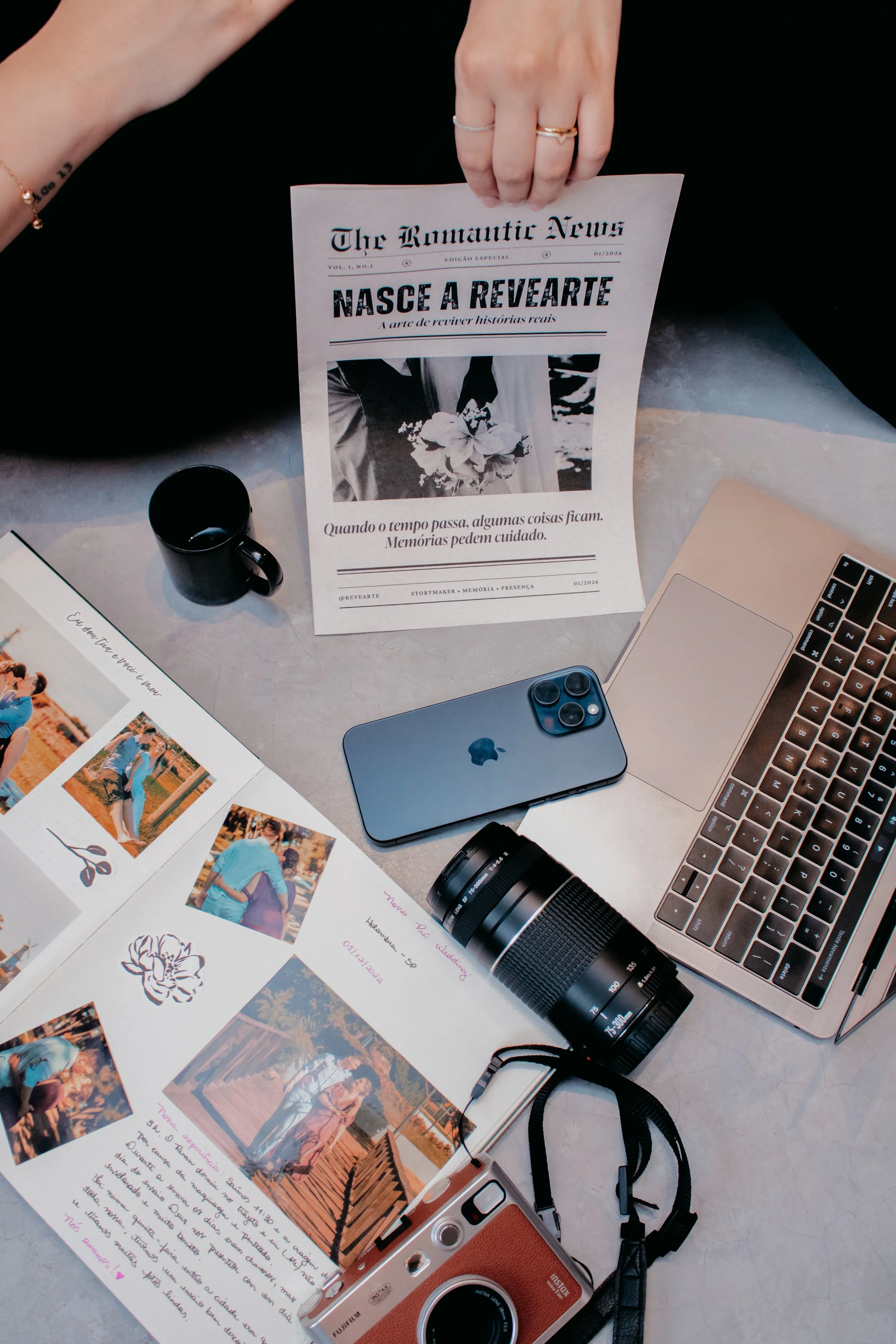 Detalhes da produção de memórias Revearte: mãos segurando jornal vintage sobre mesa de trabalho com iPhone, lente fotográfica e um álbum de fotos (scrapbook) aberto.