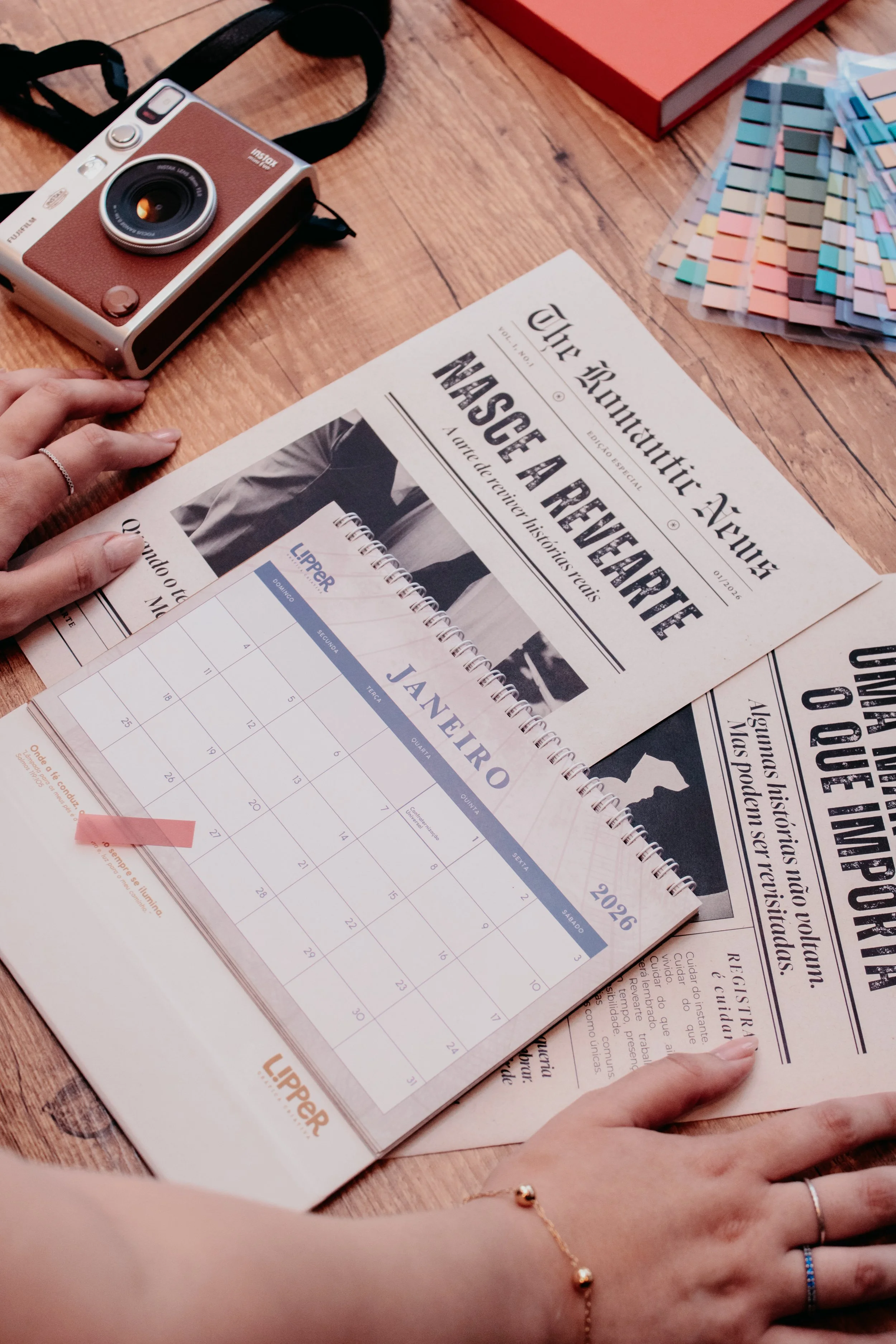 Mãos femininas organizando calendário de 2026 e jornais vintage sobre mesa de madeira com câmera fotográfica retrô.