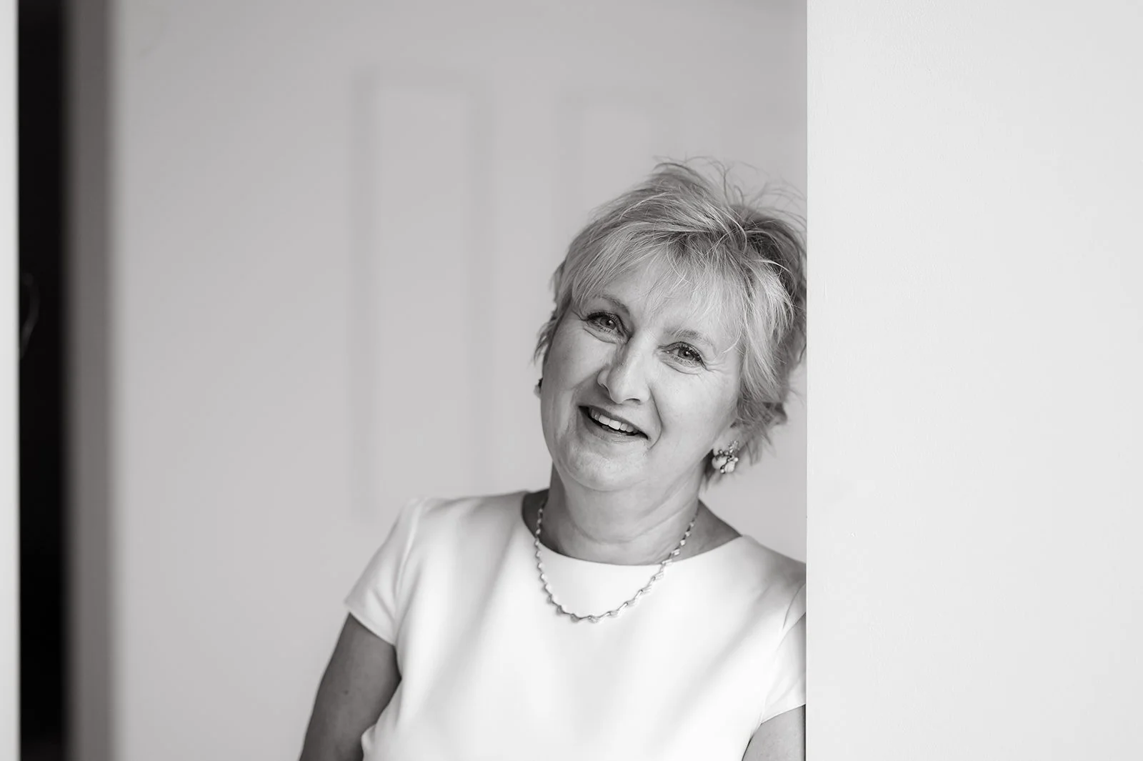 Black and white photo of a smiling woman with short, wavy hair, wearing earrings, a necklace, and a short-sleeved top, leaning against a wall.