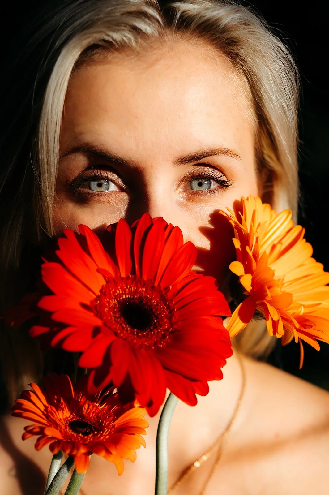A woman with blonde hair and blue eyes holding red and orange Gerbera daisies in front of her face, with one eye visible.