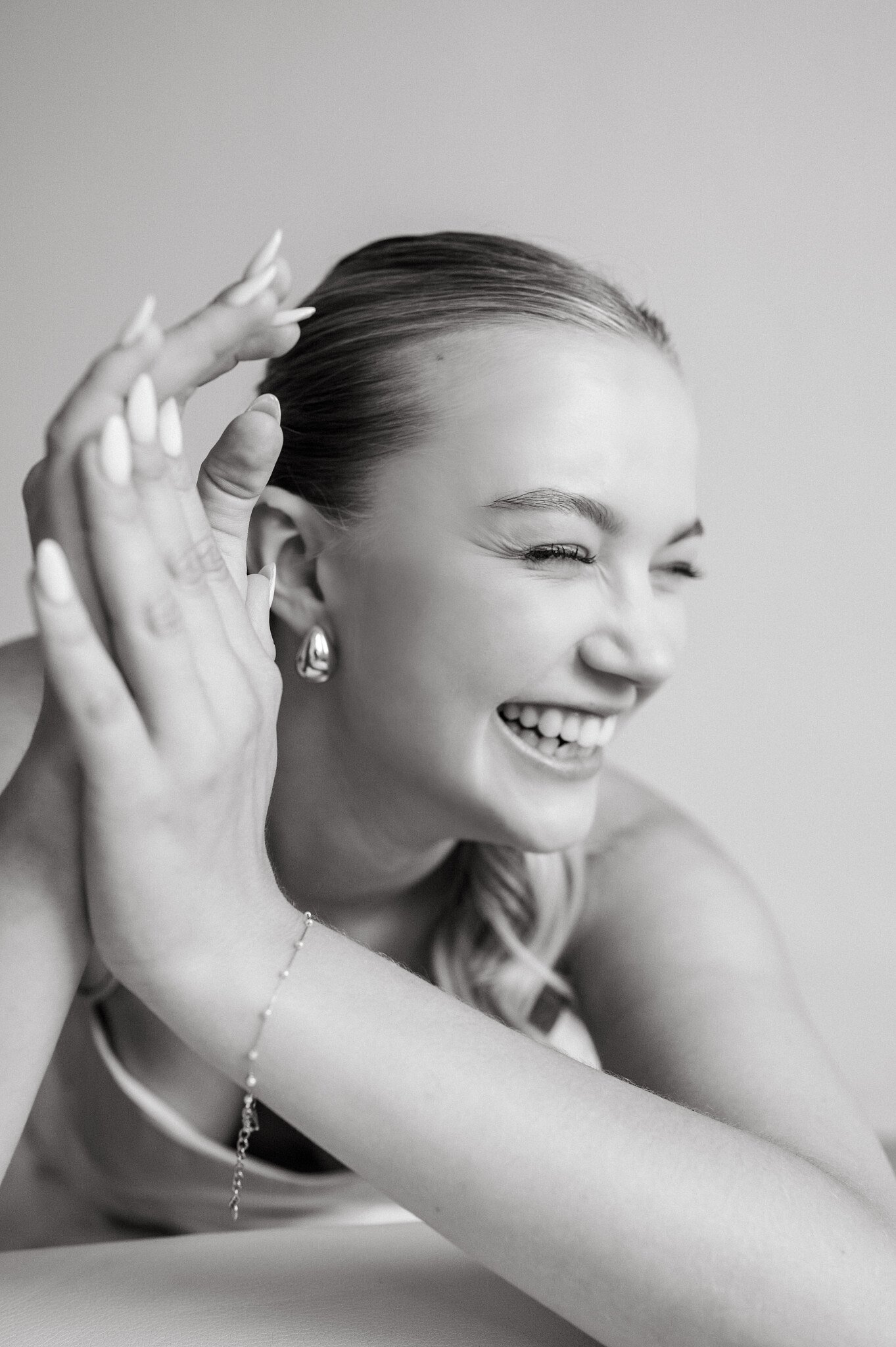 A smiling woman with light-colored lipstick, wearing earrings, a bracelet, and a tank top, posing with her head tilted and her hand near her face.