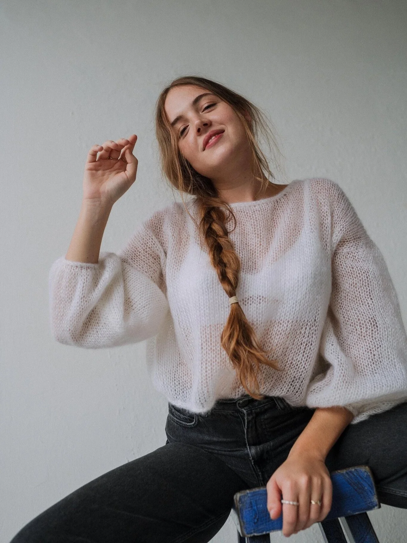 Young woman with long braided hair wearing a white knit sweater and black jeans, sitting on a stool and smiling at the camera.