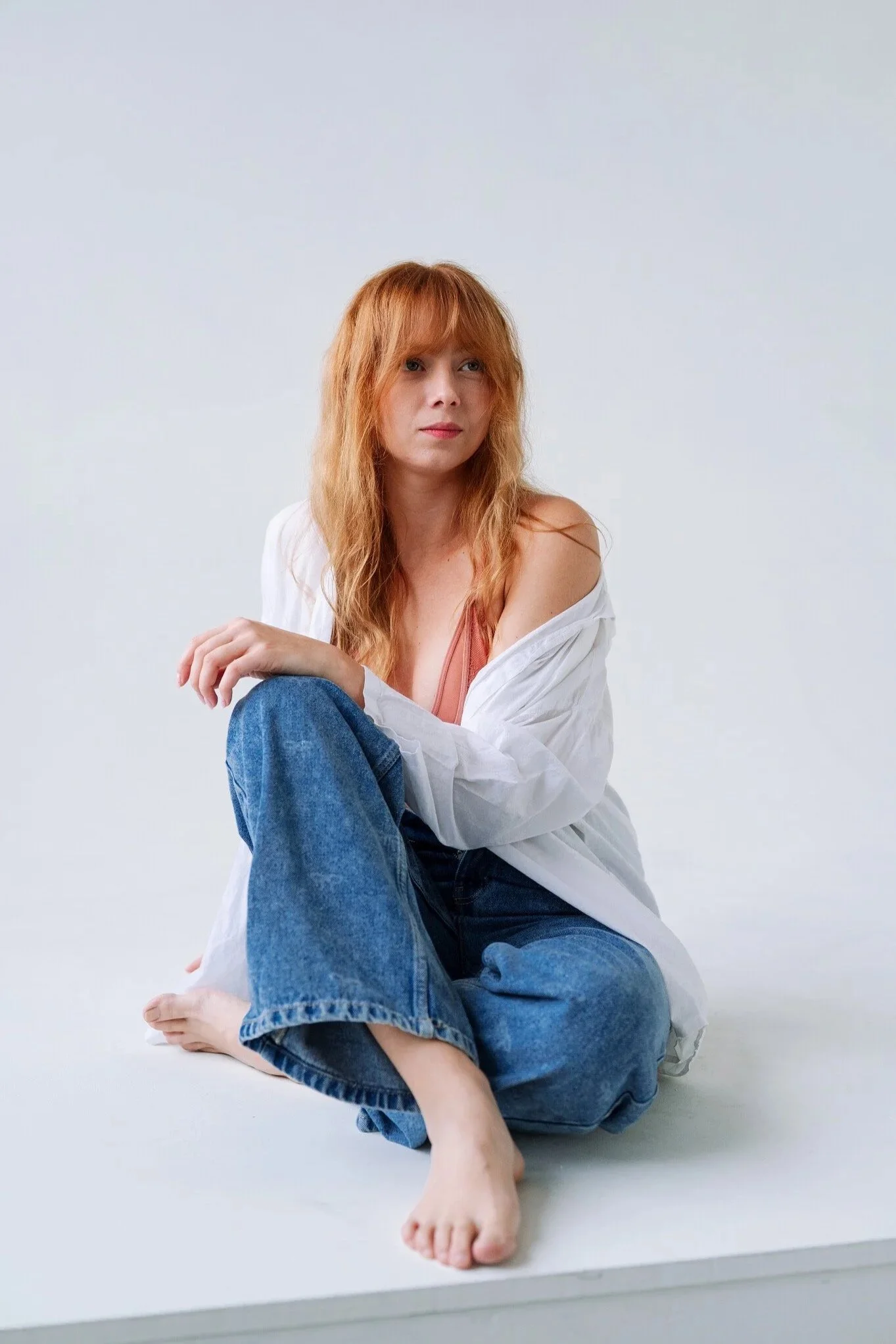 A woman with red hair and fair skin sitting on the floor in front of a plain white background, wearing a white shirt and blue jeans.