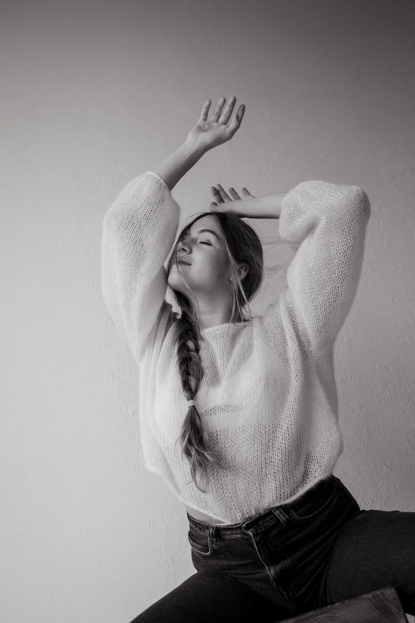 A woman with long hair in a braid, wearing a loose knit sweater and jeans, poses with her eyes closed and arms raised, against a plain wall.