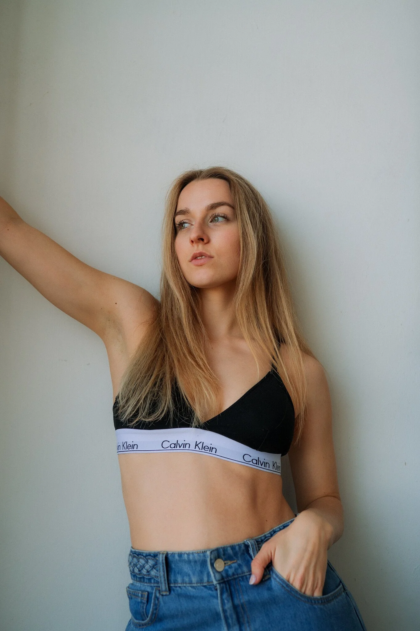 Young woman with blonde hair wearing a black Calvin Klein sports bra and blue jeans, standing against a plain white wall, looking to the side with her arm raised.