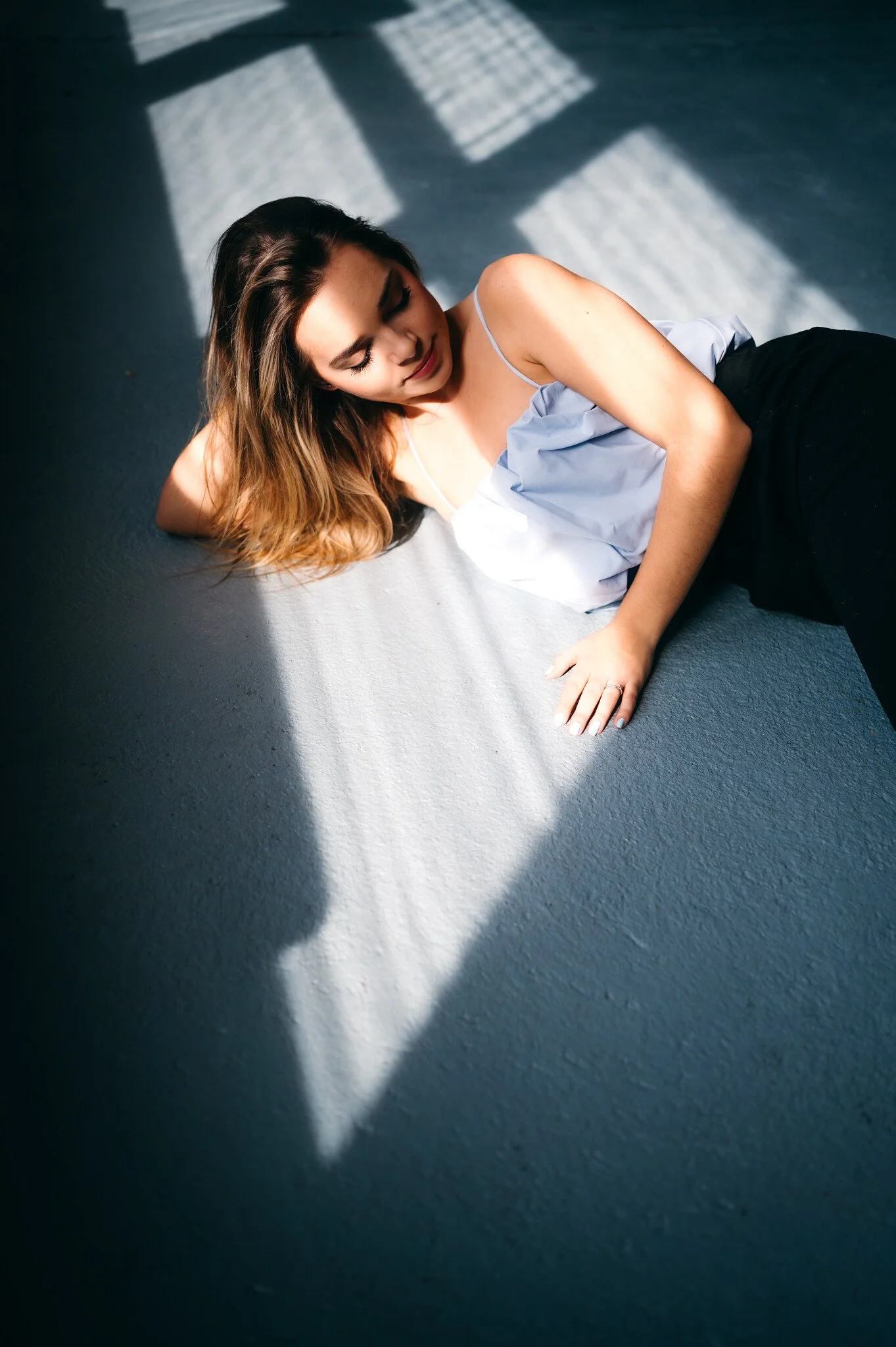 A woman lying on the floor illuminated by sunlight through window blinds, with her eyes closed and a relaxed expression.