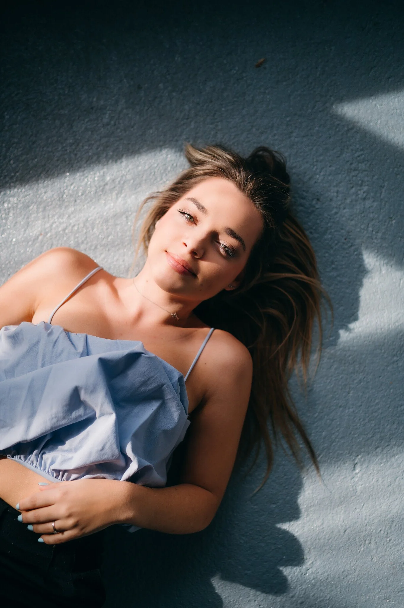 A young woman lies on the ground with her head tilted slightly, looking at the camera with a relaxed expression. She has long, brown hair spread out around her, and is wearing a light blue, thin-strapped top. The ground appears to be a textured, gray