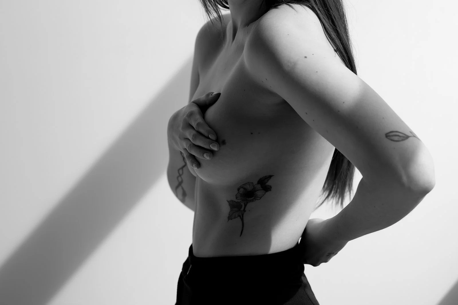 Black and white photo of a topless woman with tattoos covering her side, arm, and hand, standing against a plain white background.
