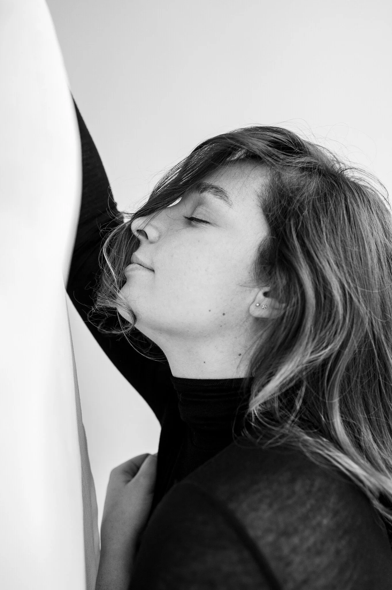 Black and white photo of a young woman resting her face against a wall with closed eyes, wearing a black top and earrings.
