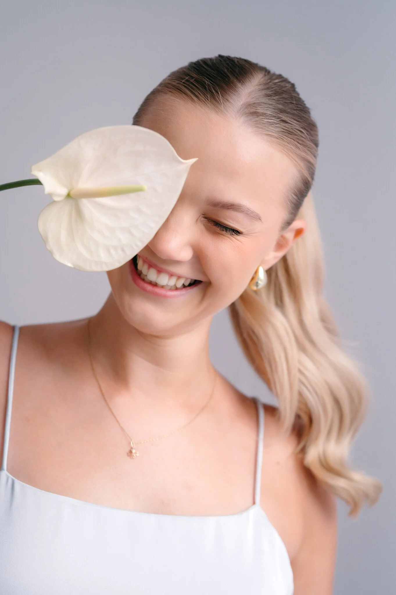 A smiling woman with blonde hair tied in a ponytail holds a white calla lily flower close to her face, covering one eye, against a plain background.