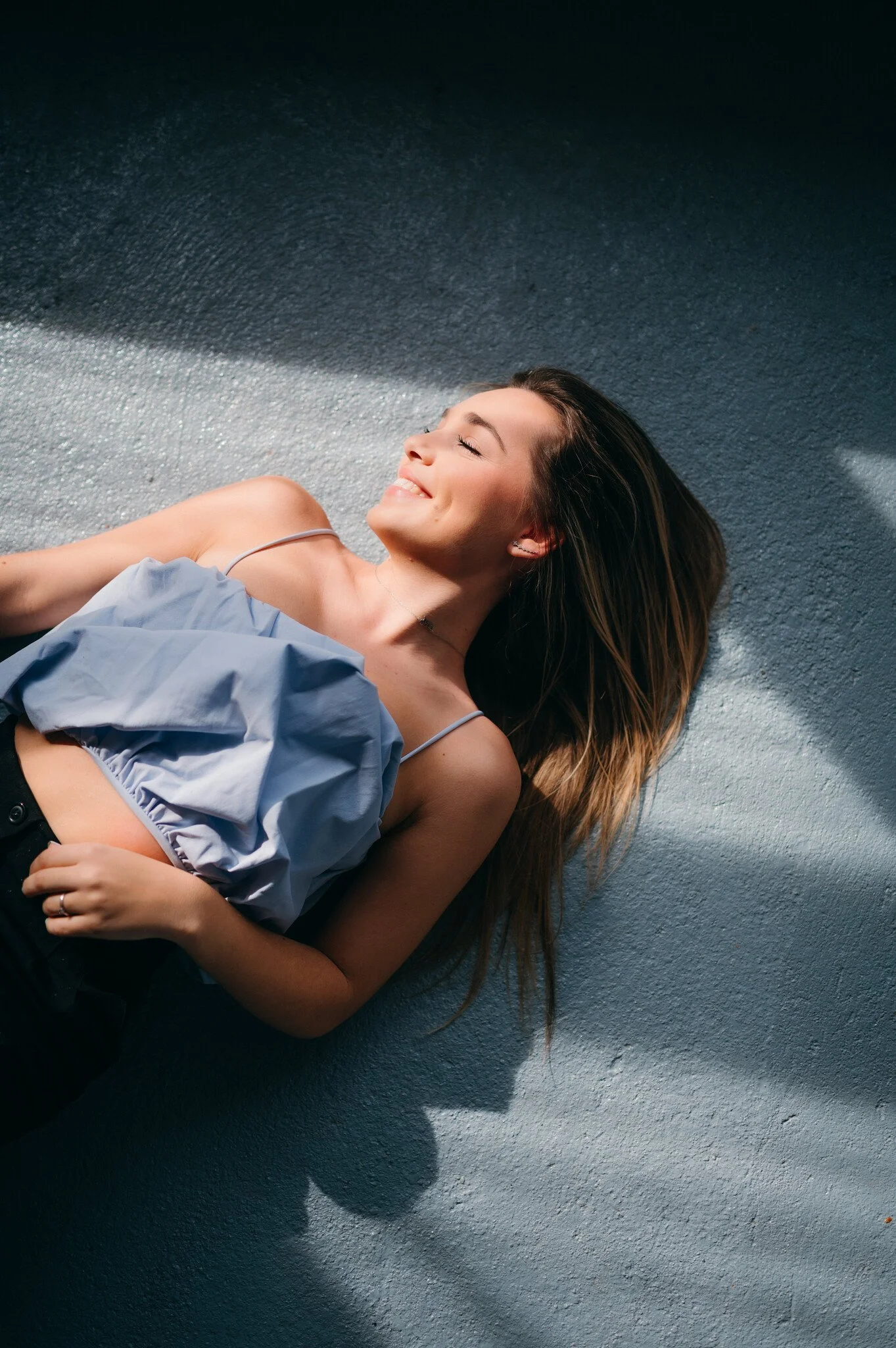 A woman lying on textured concrete ground with sunlight and shadows, smiling with eyes closed, wearing a light blue sleeveless top and black pants.