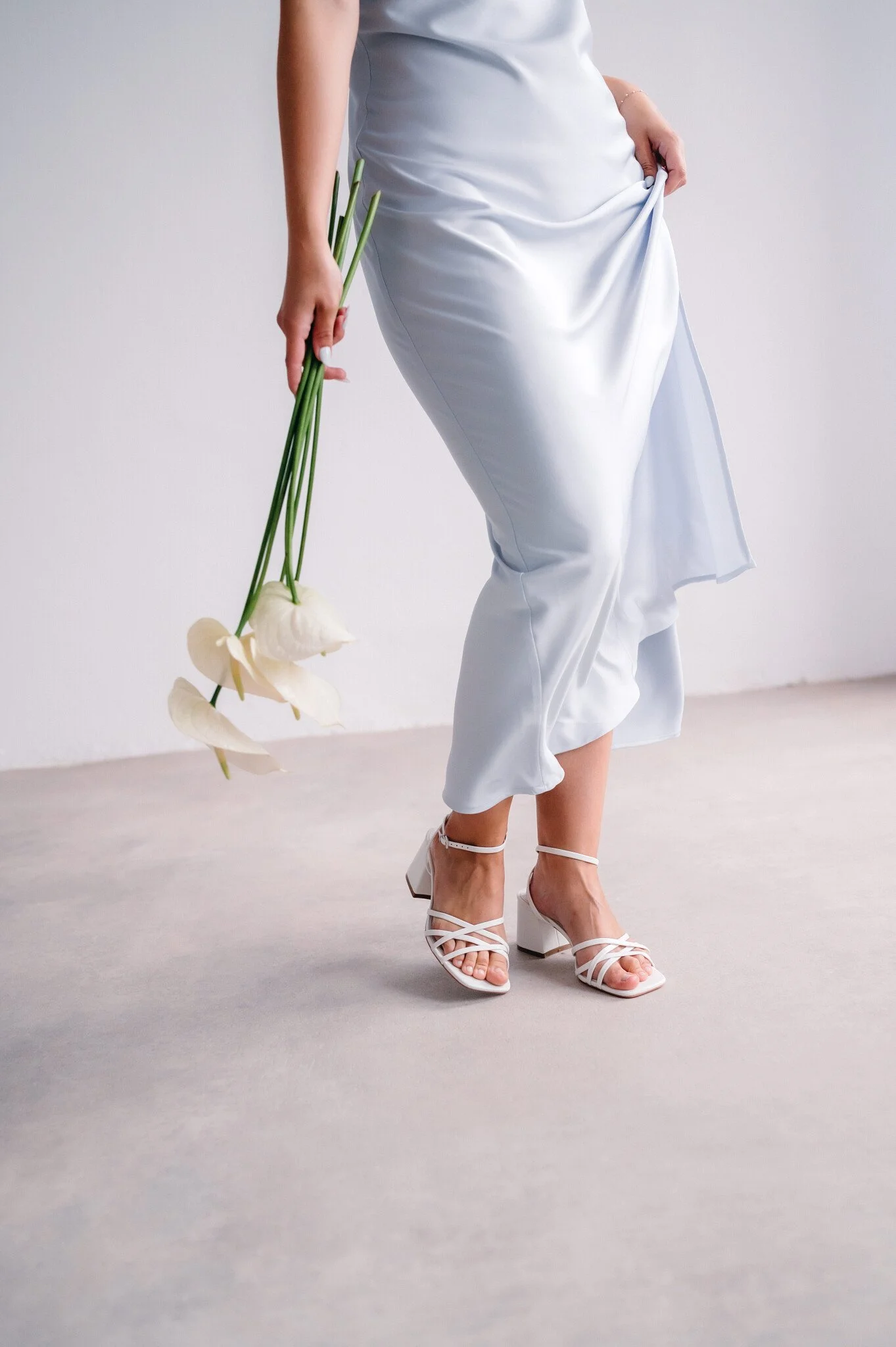 A woman wearing a white satin dress and white heeled sandals holding three white calla lilies.