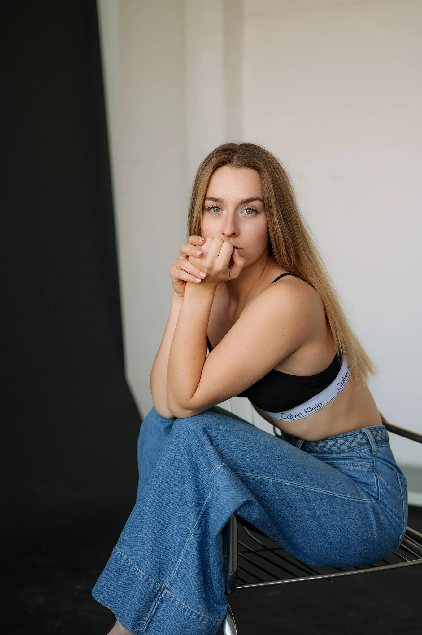 A young woman with long, light brown hair sitting sideways on a metal bench, wearing a black Calvin Klein sports bra and blue jeans, with her hands clasped near her face, looking directly at the camera against a neutral background.