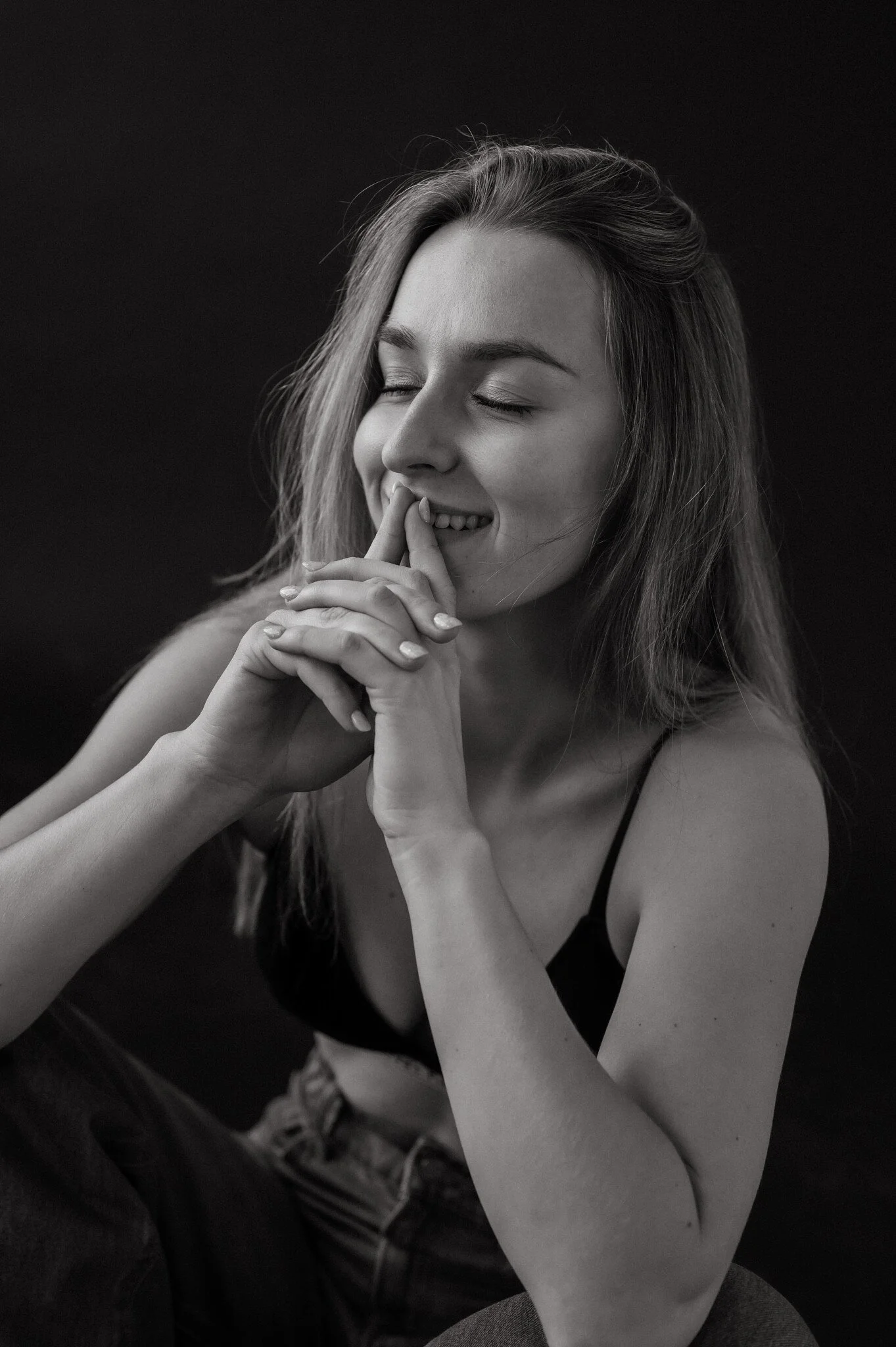 Black and white photo of a young woman smiling with her eyes closed, hands gently placed near her face, wearing a spaghetti strap top.
