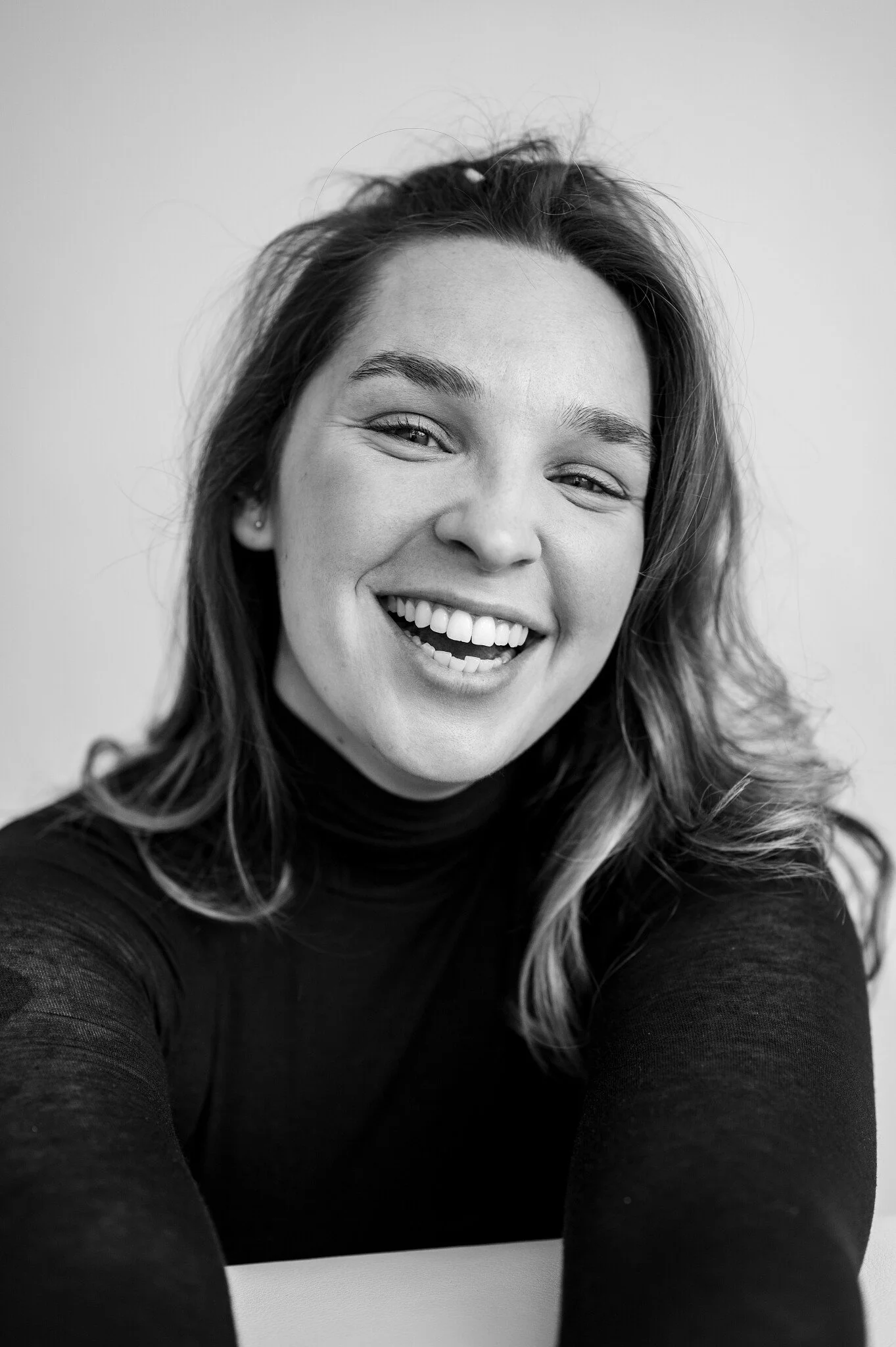 A woman with wavy hair, wearing a black turtleneck sweater, smiling and taking a selfie.