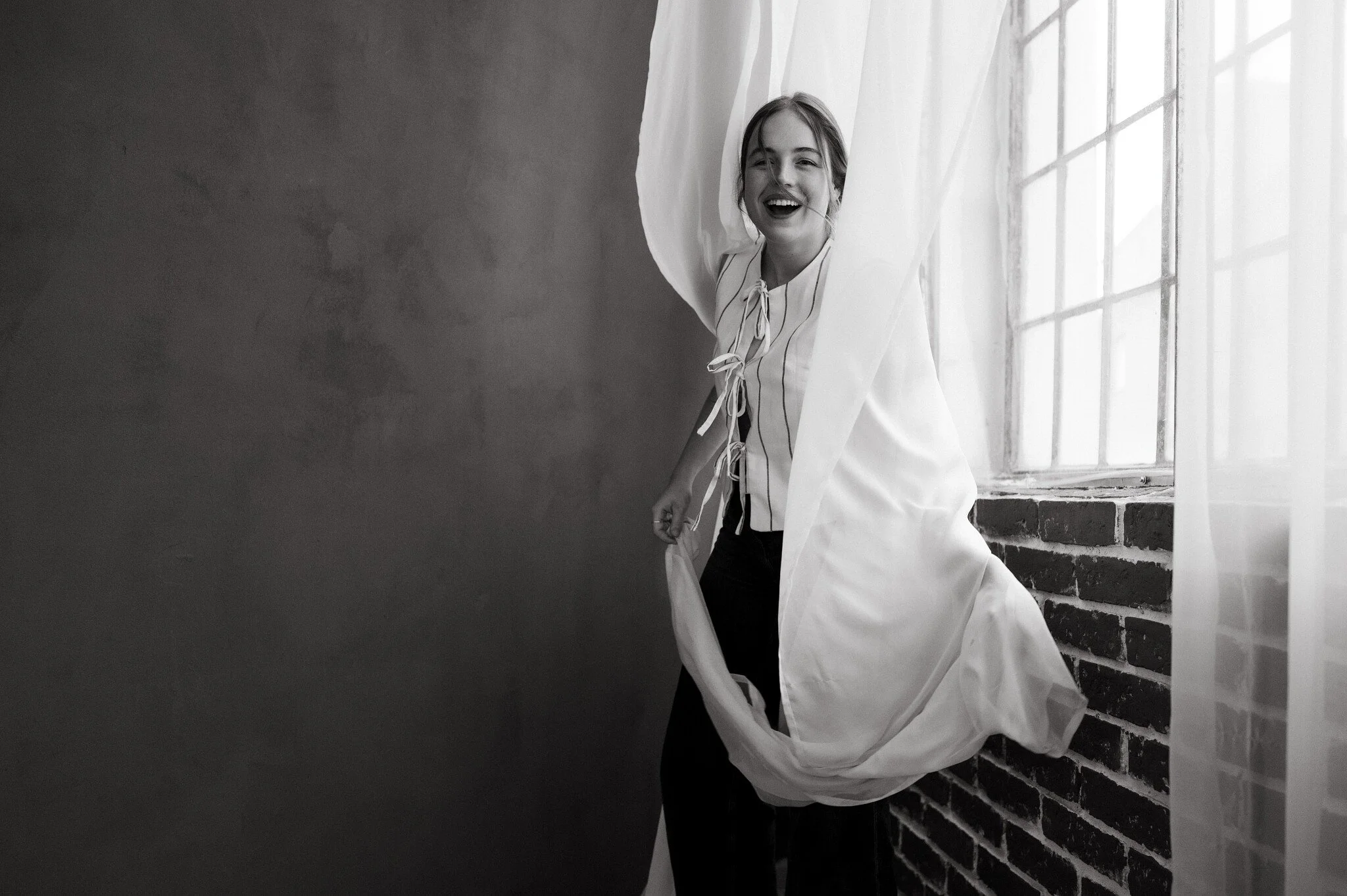 A woman smiling and curtain flowing near a brick wall by a large window in black and white.