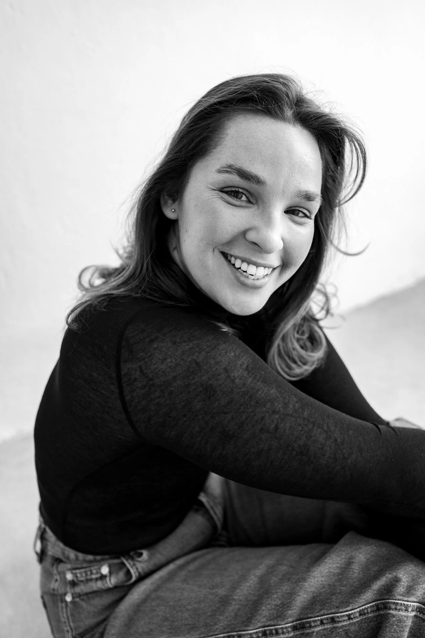 Black and white portrait of a young woman smiling, sitting with her arms crossed and wearing a long-sleeve shirt and jeans.