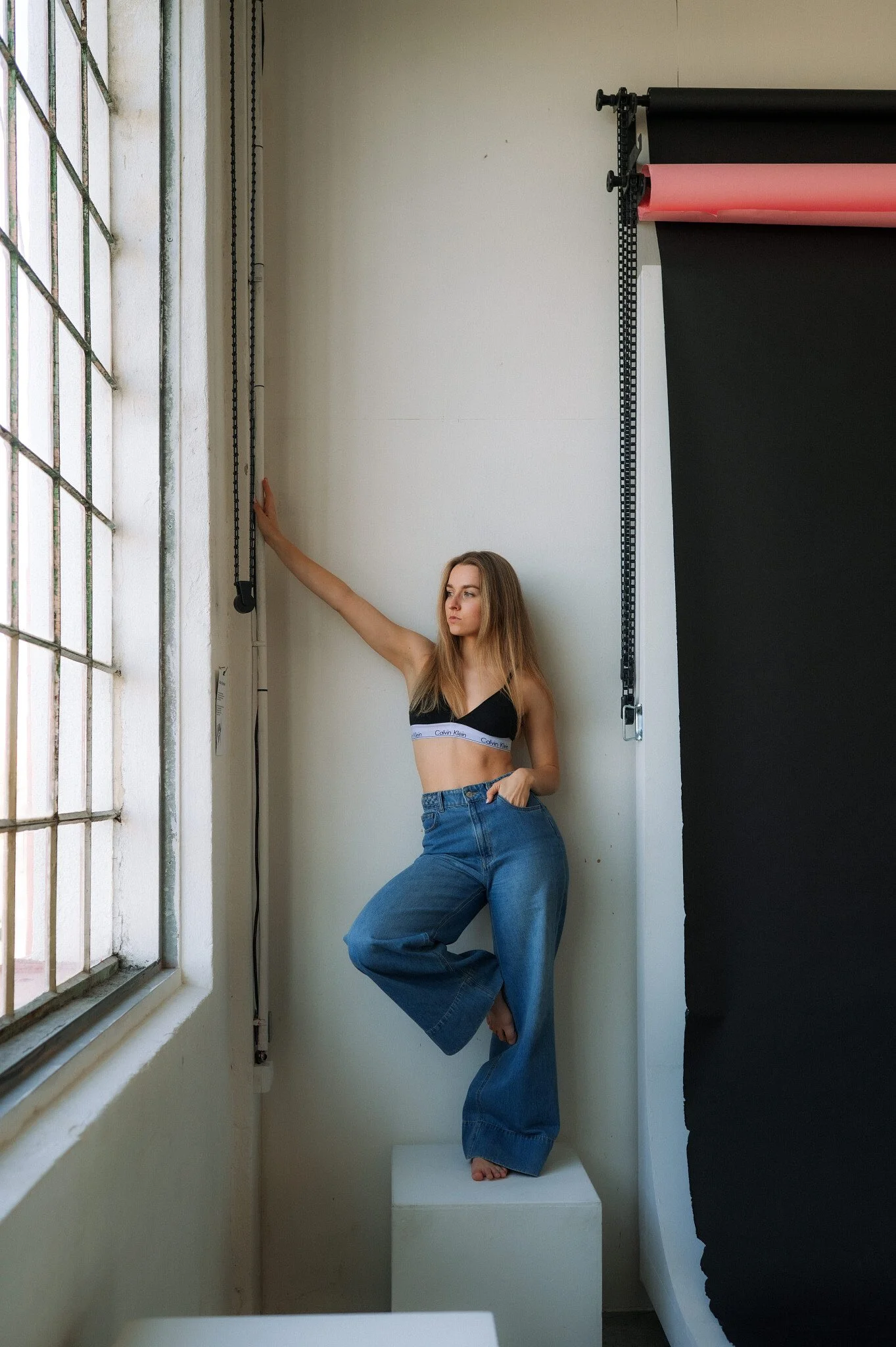 A young woman with long blond hair in casual clothing, standing on a white rectangular block in a photography studio, holding a chain on the wall, next to a large window with metal grid and a black photo backdrop.