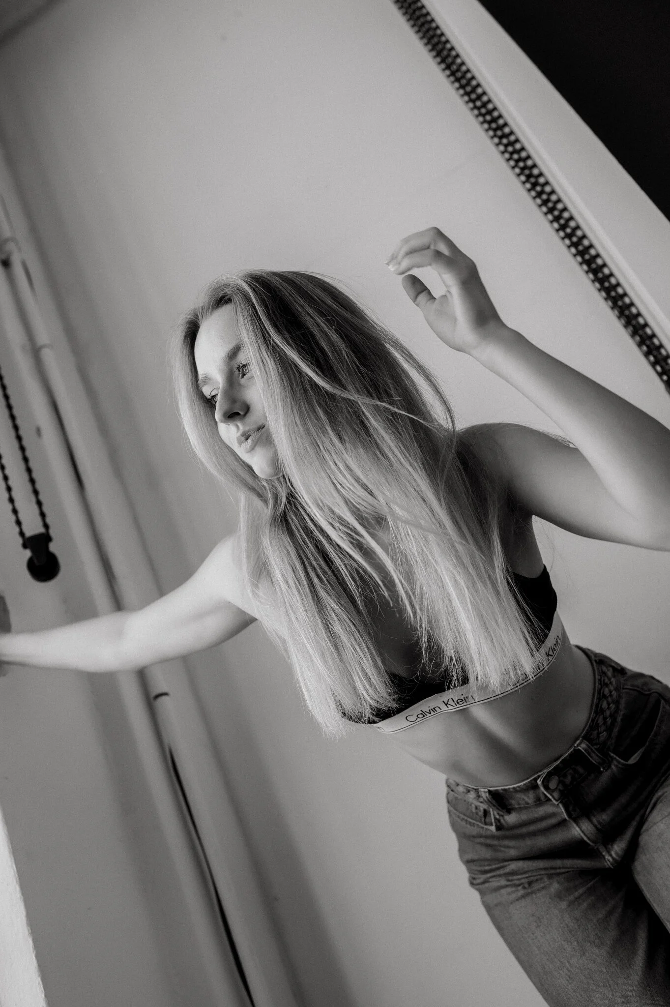 Black and white photo of a woman with long, straight hair, wearing a Calvin Klein sports bra and jeans, standing in front of a mirror with her hand raised.