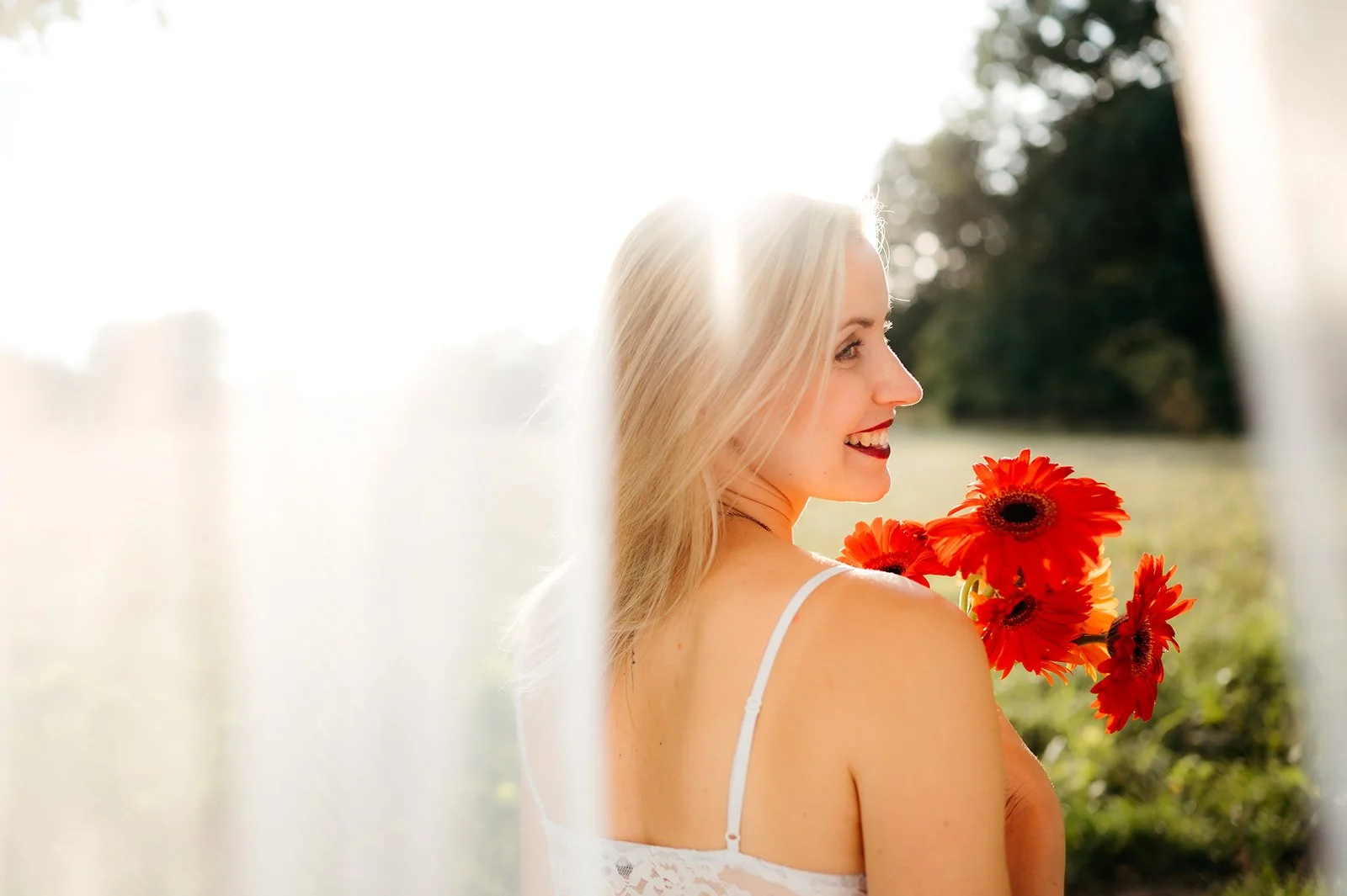 A blonde woman in a white dress holding bright orange flowers, smiling outdoors with sunlight in the background.