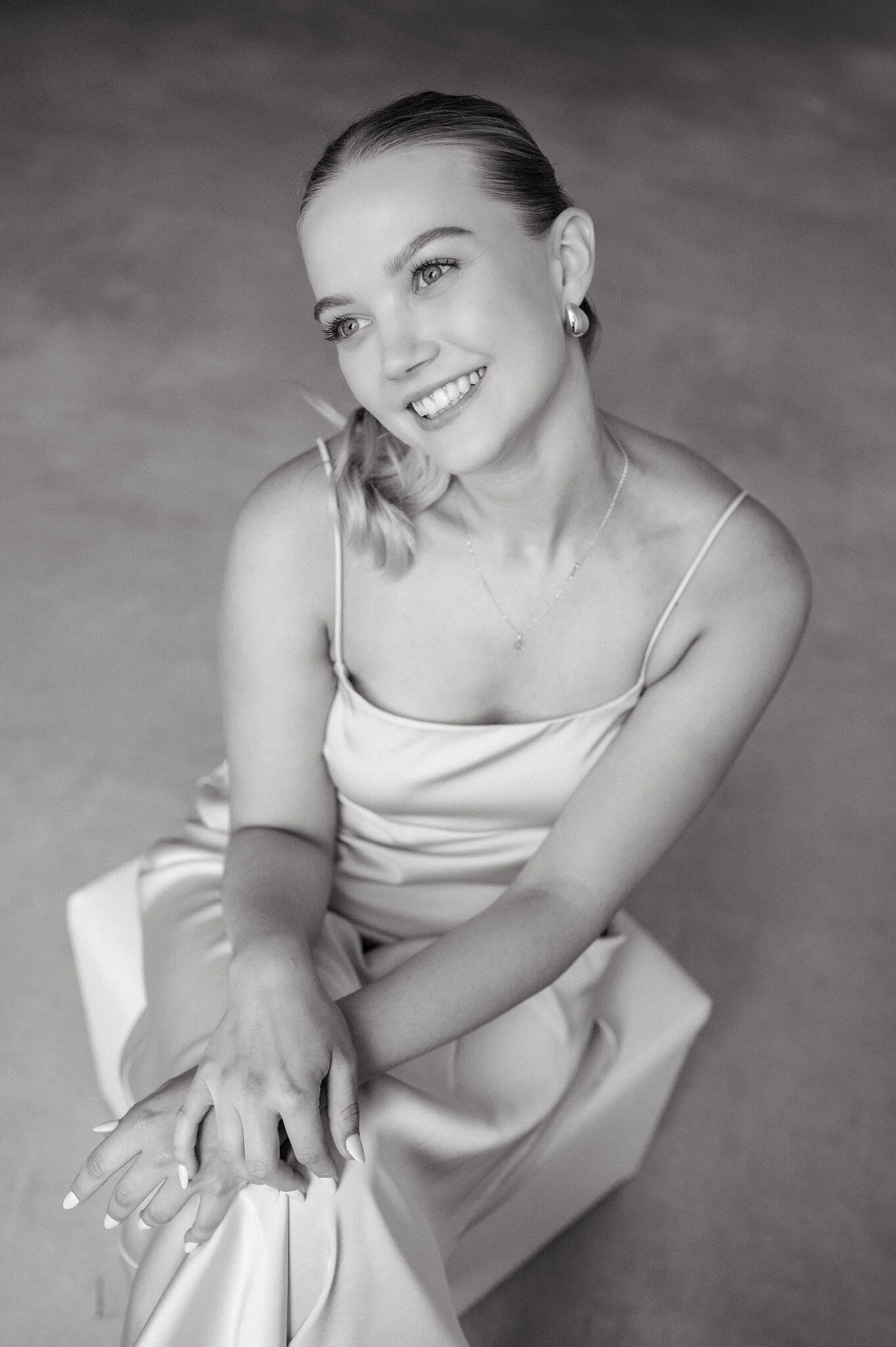 Black and white portrait of a smiling woman sitting on a box, wearing a silk dress with thin straps, looking slightly up and to the side.