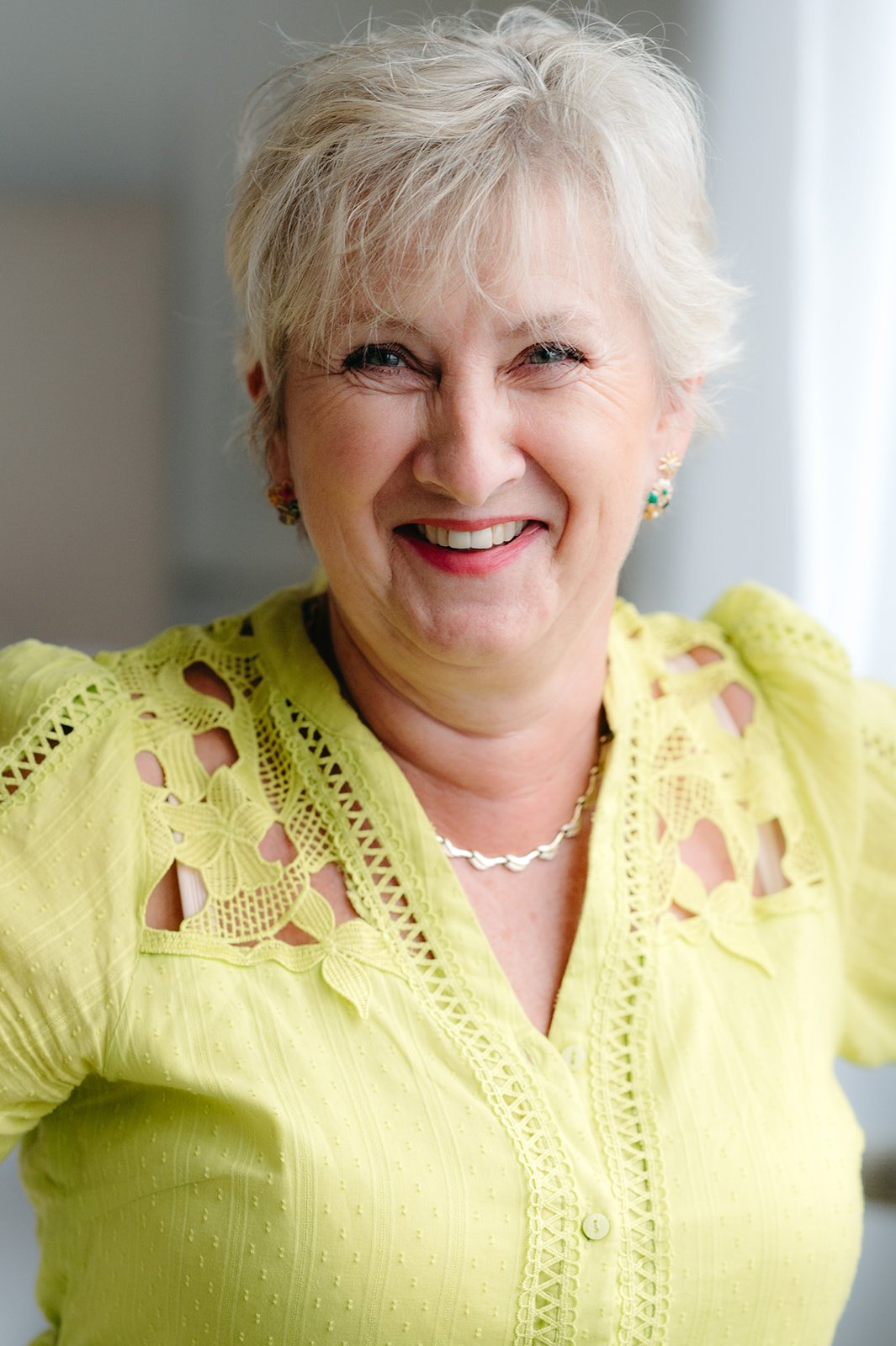 A smiling woman with short, gray hair wearing a yellow, embroidered blouse and a silver necklace.