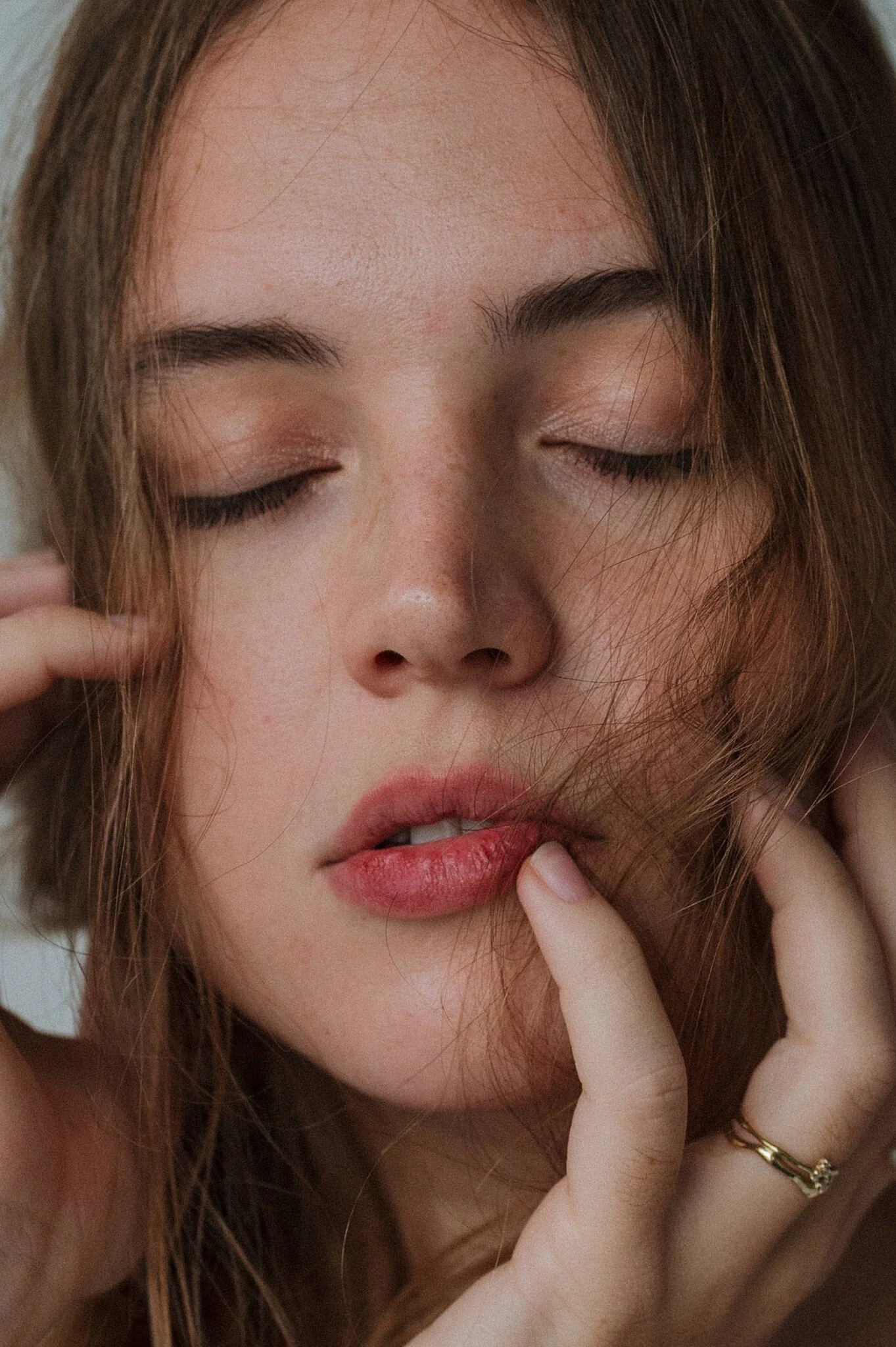 Close-up of a young woman with closed eyes, touching her face and lips, with loose hair partially covering her face.
