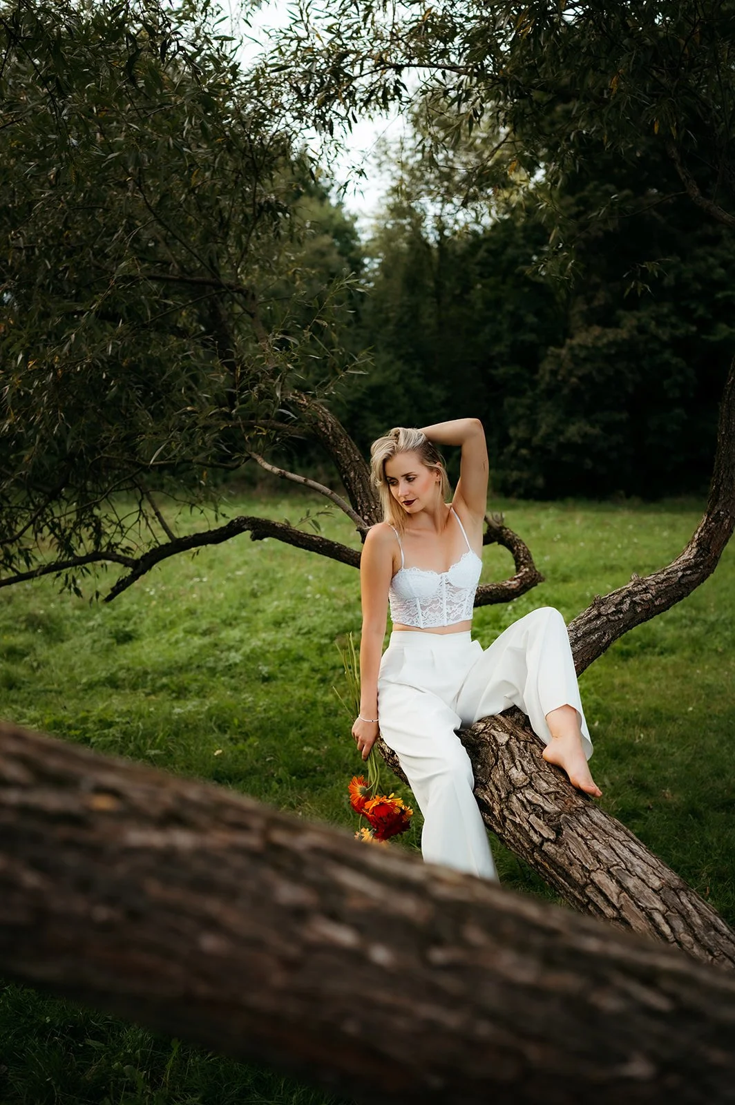 A woman sitting on a tree branch in a grassy outdoor setting, wearing white clothing and holding a bouquet of orange flowers.