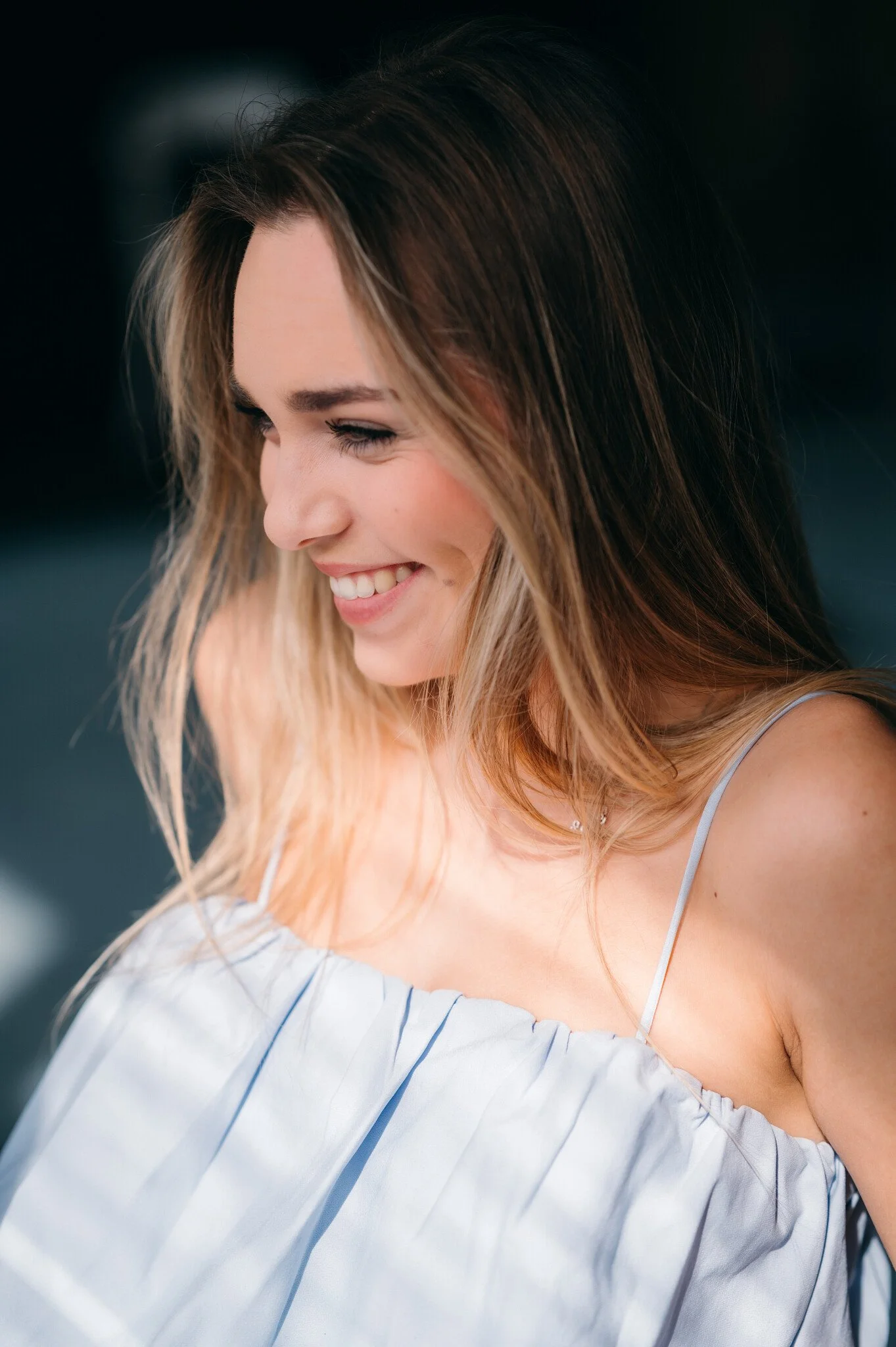 Close-up of a young woman with brown hair smiling and looking down. She is wearing a light-colored, off-the-shoulder top.