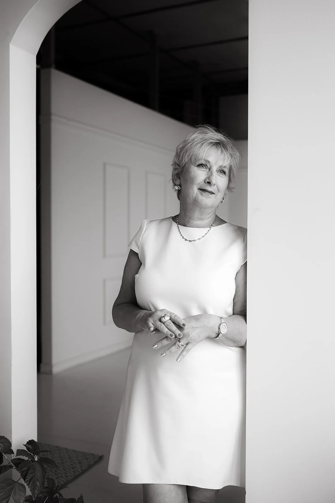 A senior woman in a white dress standing in a doorway, looking outside with a slight smile.