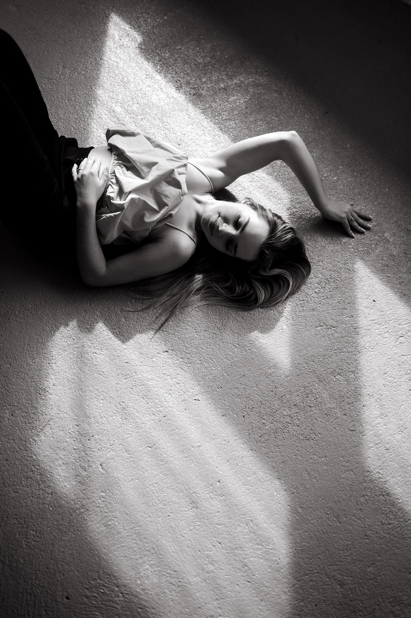 A young woman lying on the floor in black and white, with her eyes closed and her hand resting on her stomach, illuminated by sunlight creating a shadow on the textured surface.