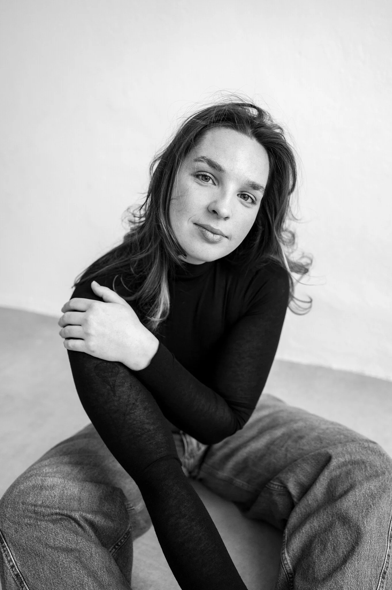 Black and white portrait of a young woman with wavy hair, wearing a black long-sleeve shirt and jeans, sitting on the floor with one arm crossed over her chest, looking at the camera.