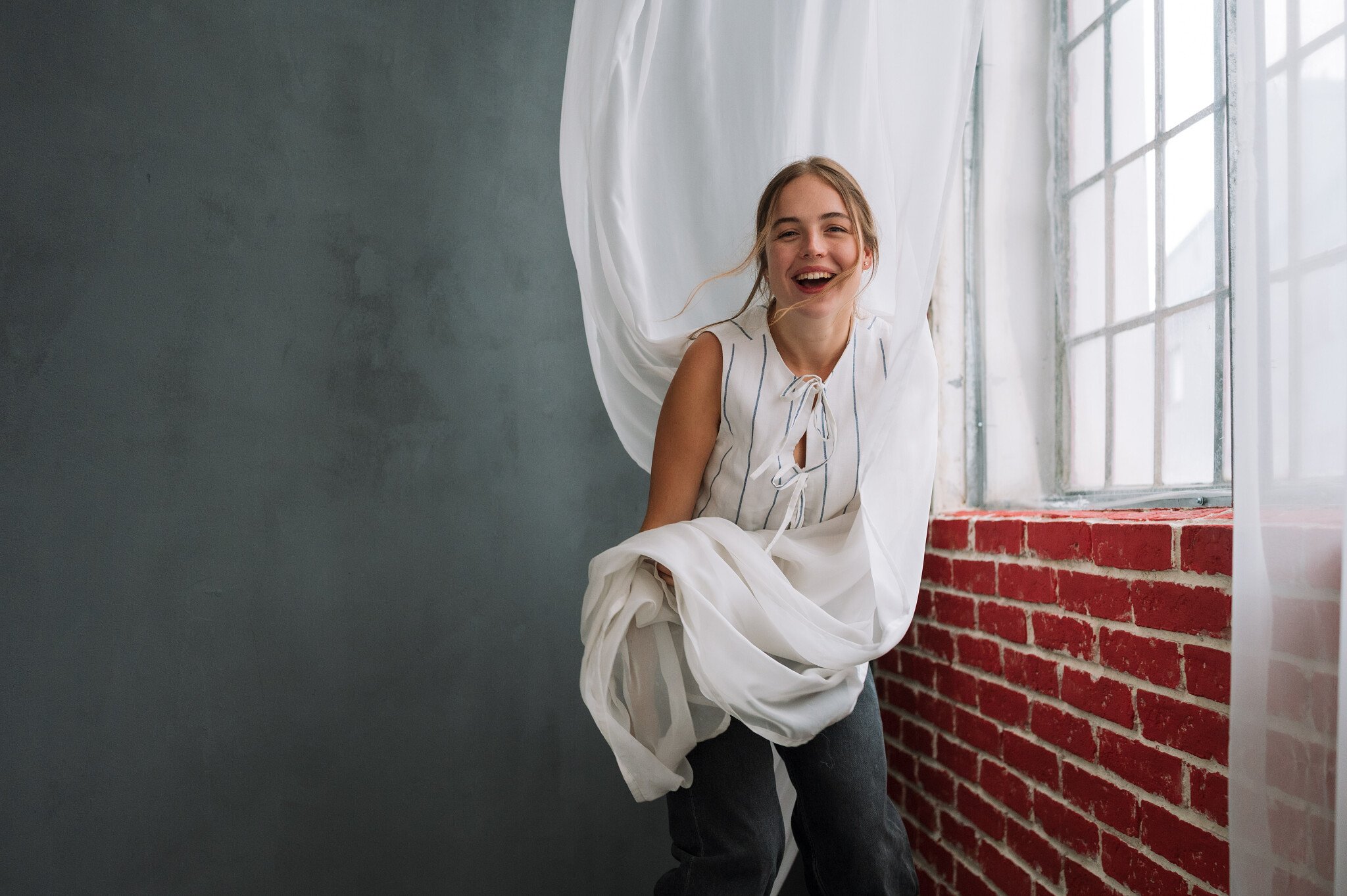 A young woman smiling and leaning out from behind white curtains near a large window with red brick wall underneath.
