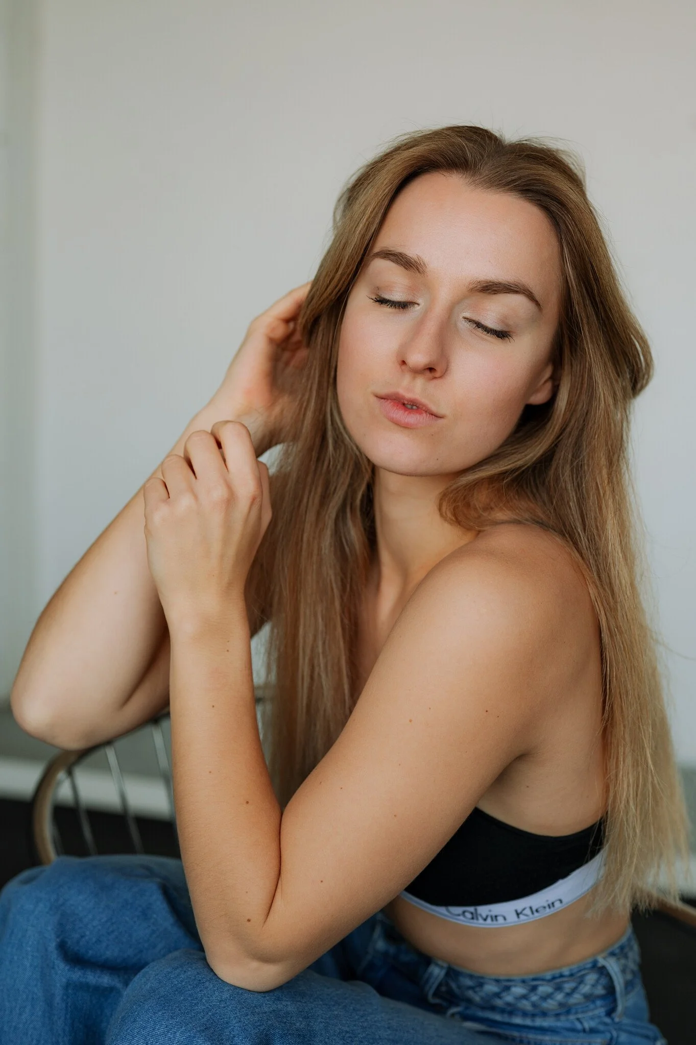 A young woman with long, light brown hair, closed eyes, and a calm expression, sitting with her arms crossed and touching her hair.