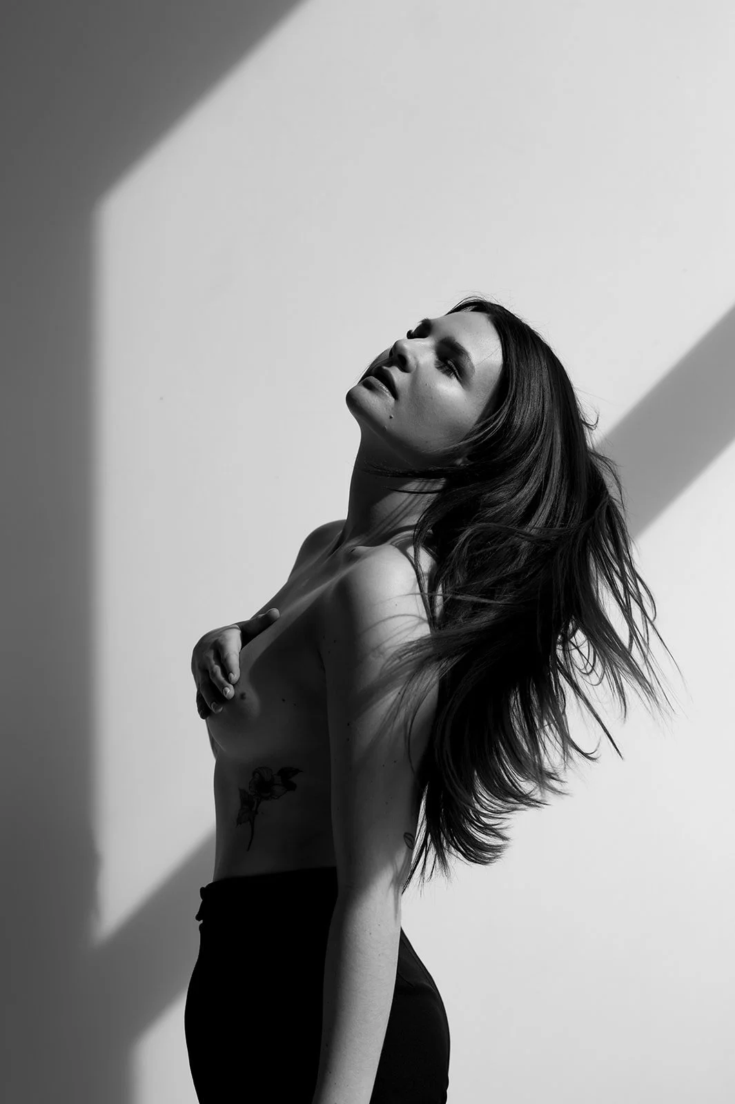 Black and white photograph of a topless woman with long hair posing against a plain background with dramatic lighting.