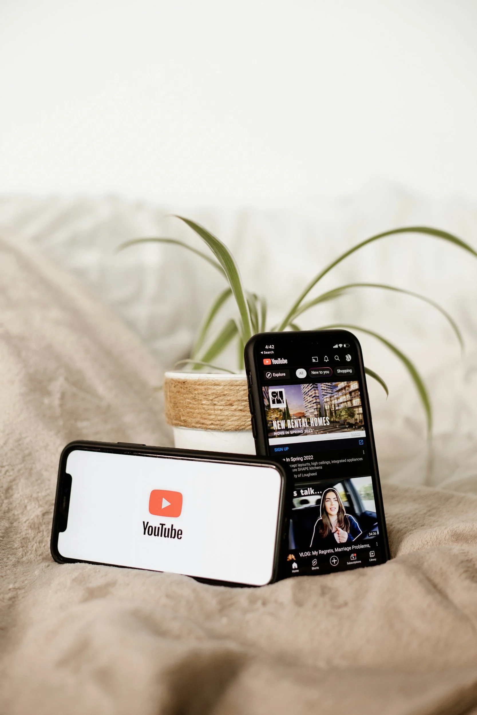 Two smartphones displaying YouTube app screens on a beige fabric surface, with a potted plant in the background.