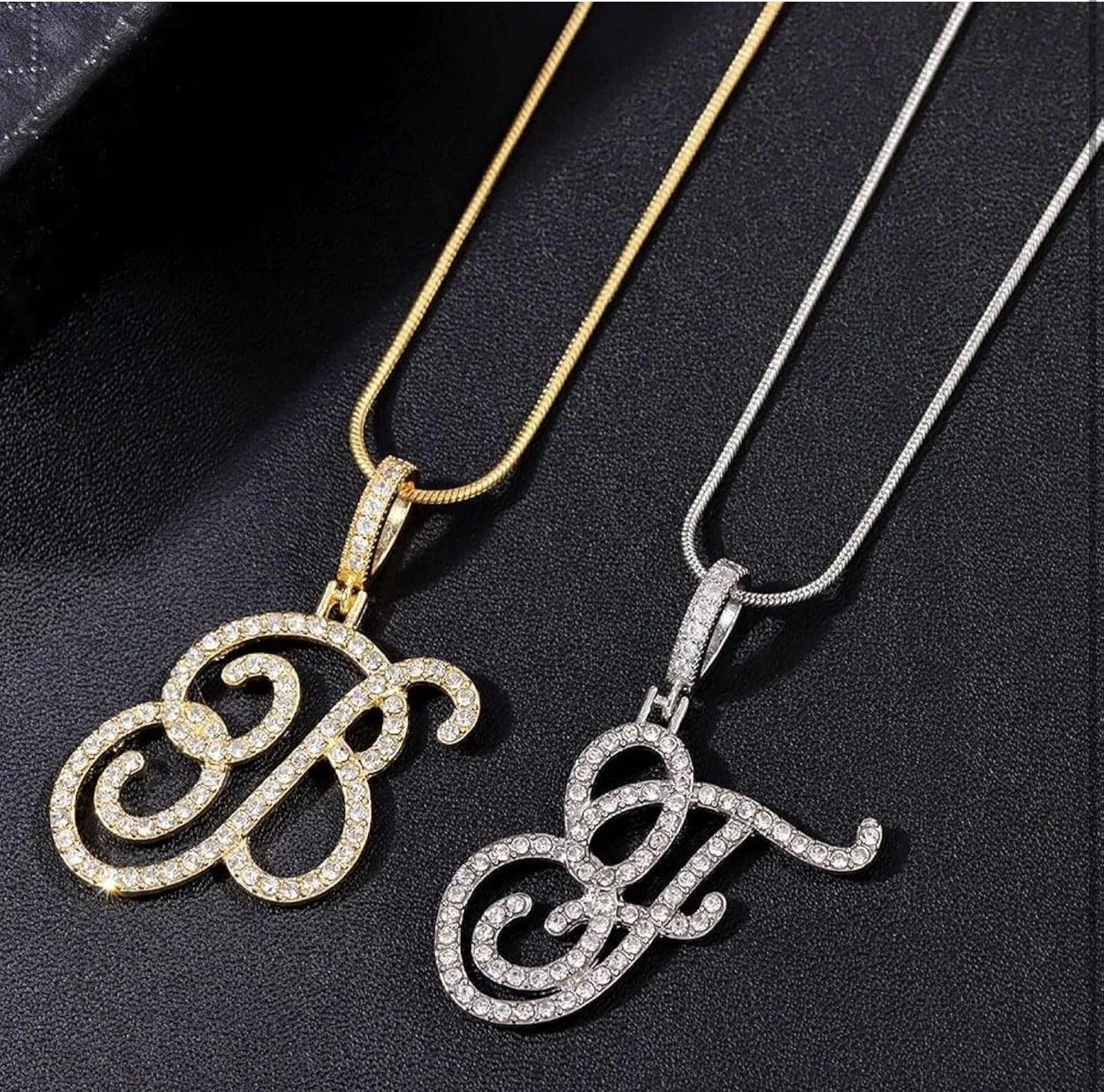 Two necklaces with pendant charms spelling out initials, one in gold with rhinestones and the other in silver with rhinestones, on black textured background.