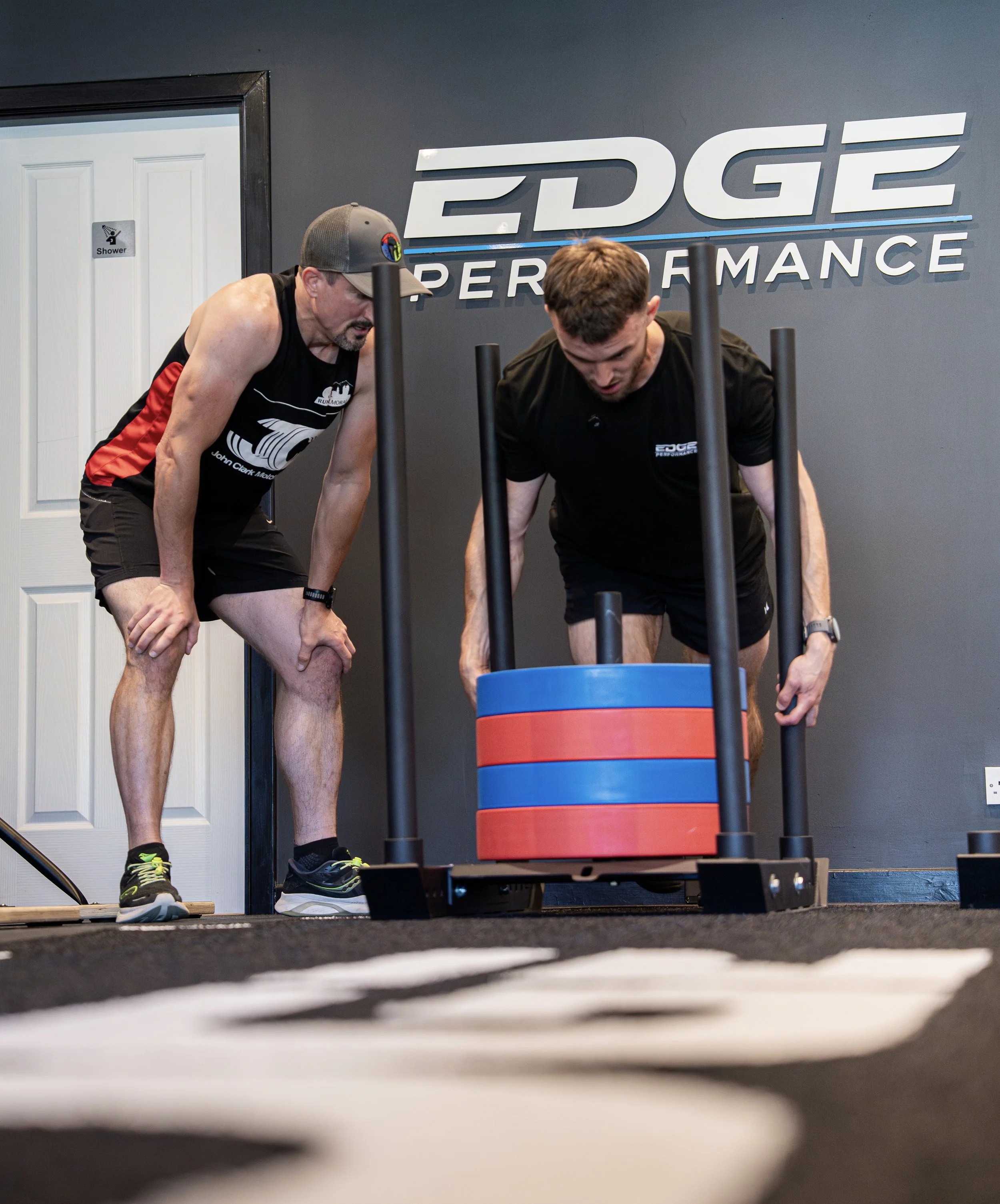 Two men working out at a gym with a sled, one pushing it while the other coaches. The wall behind has a sign that says 'EDGE PERFORMANCE'.