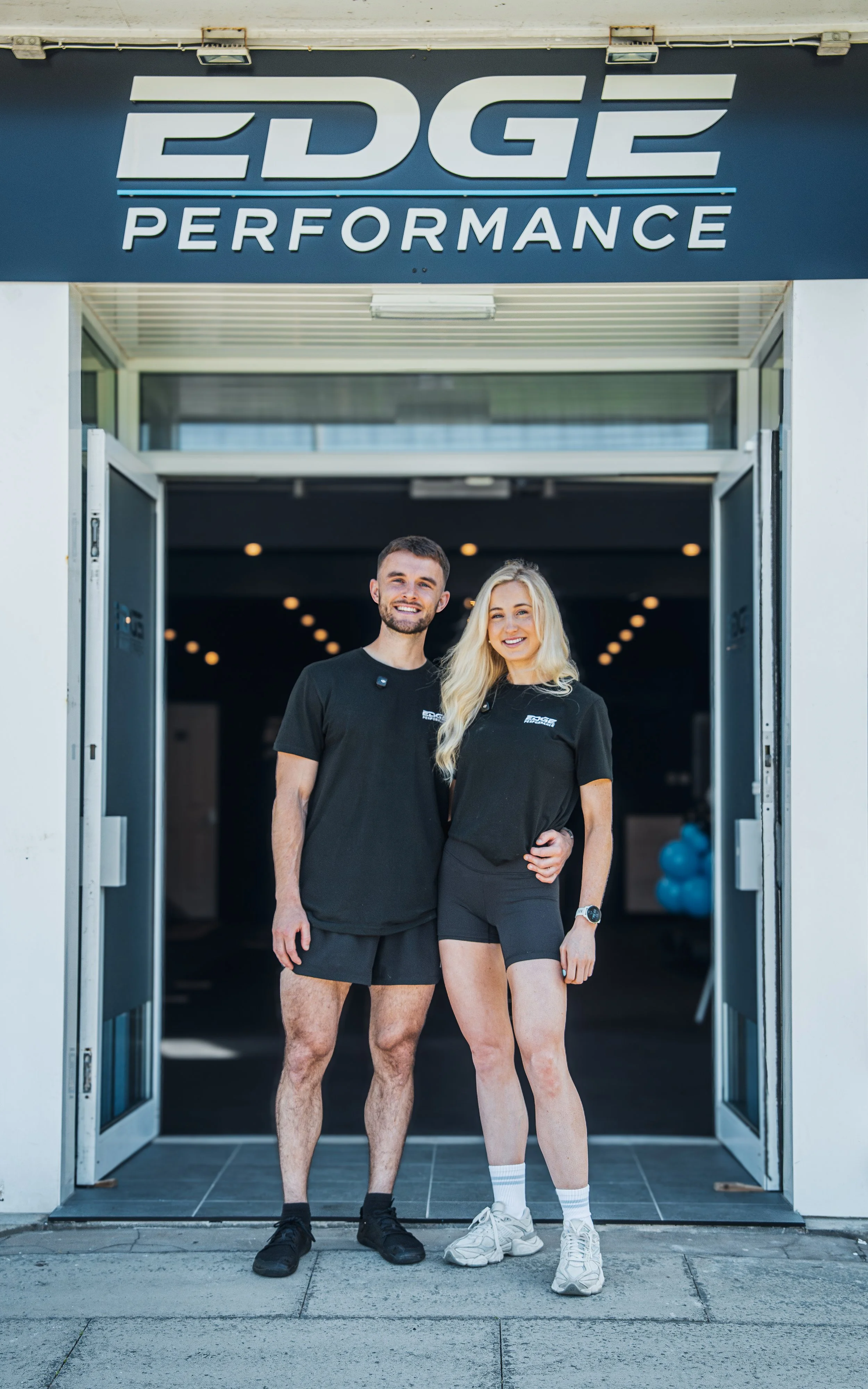 Two smiling fitness trainers, a man and a woman, standing outside the entrance of Edge Performance gym, wearing black workout shirts and shorts, with the gym sign above.