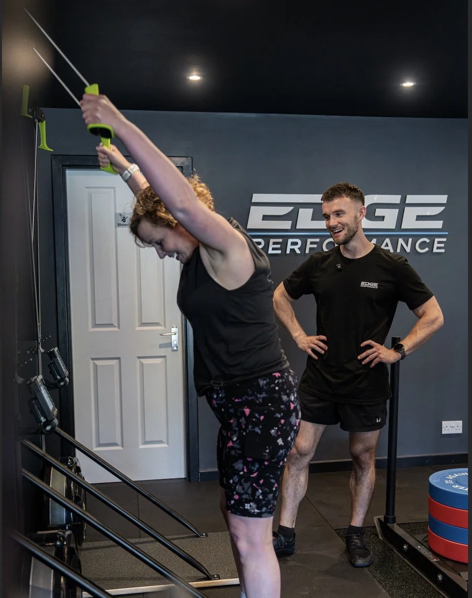 Woman exercising with a resistance band in a gym while a trainer watches and smiles.
