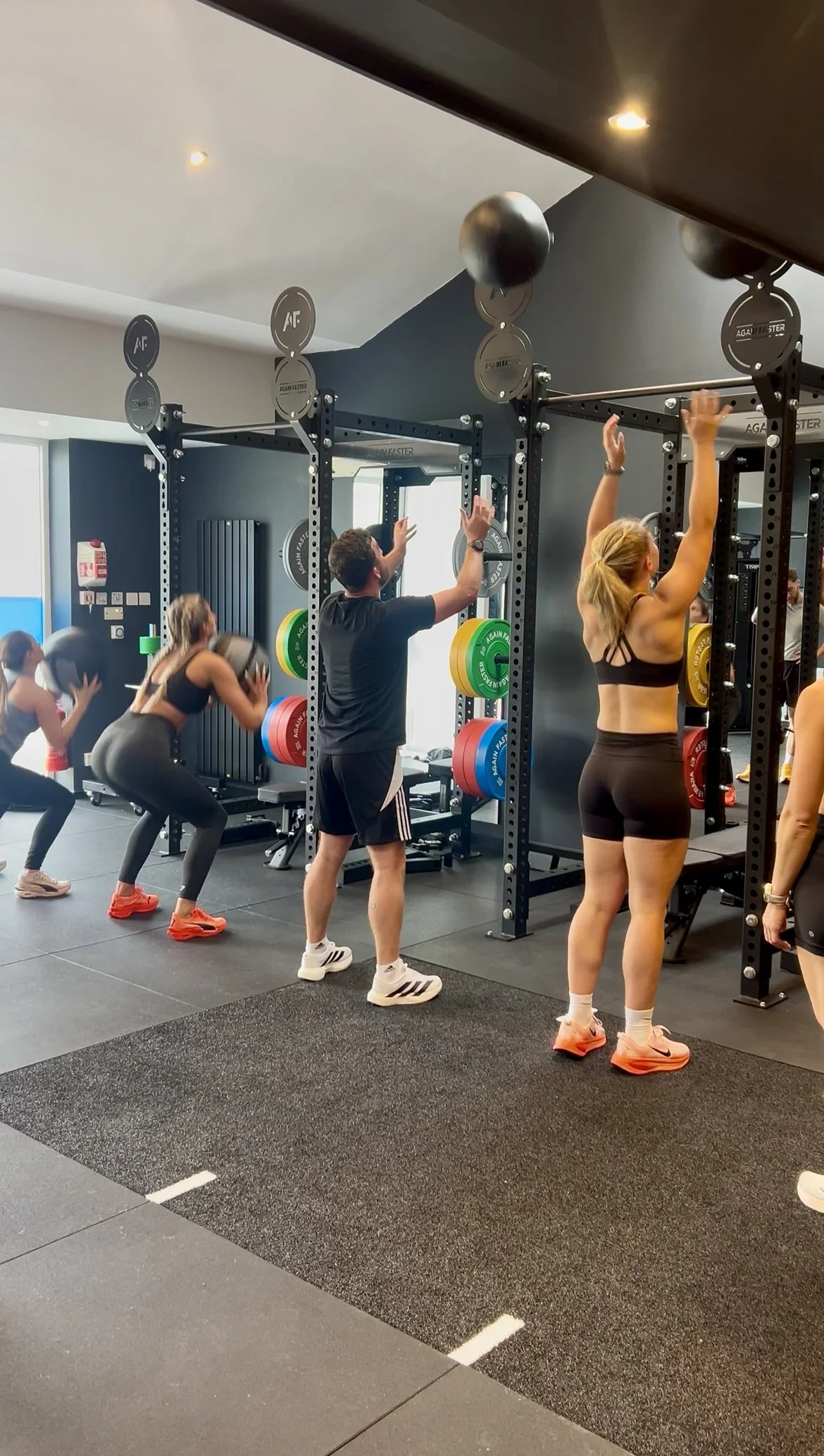 People exercising in a gym, including women lifting weights and a man standing with arms raised, with various fitness equipment in the background.