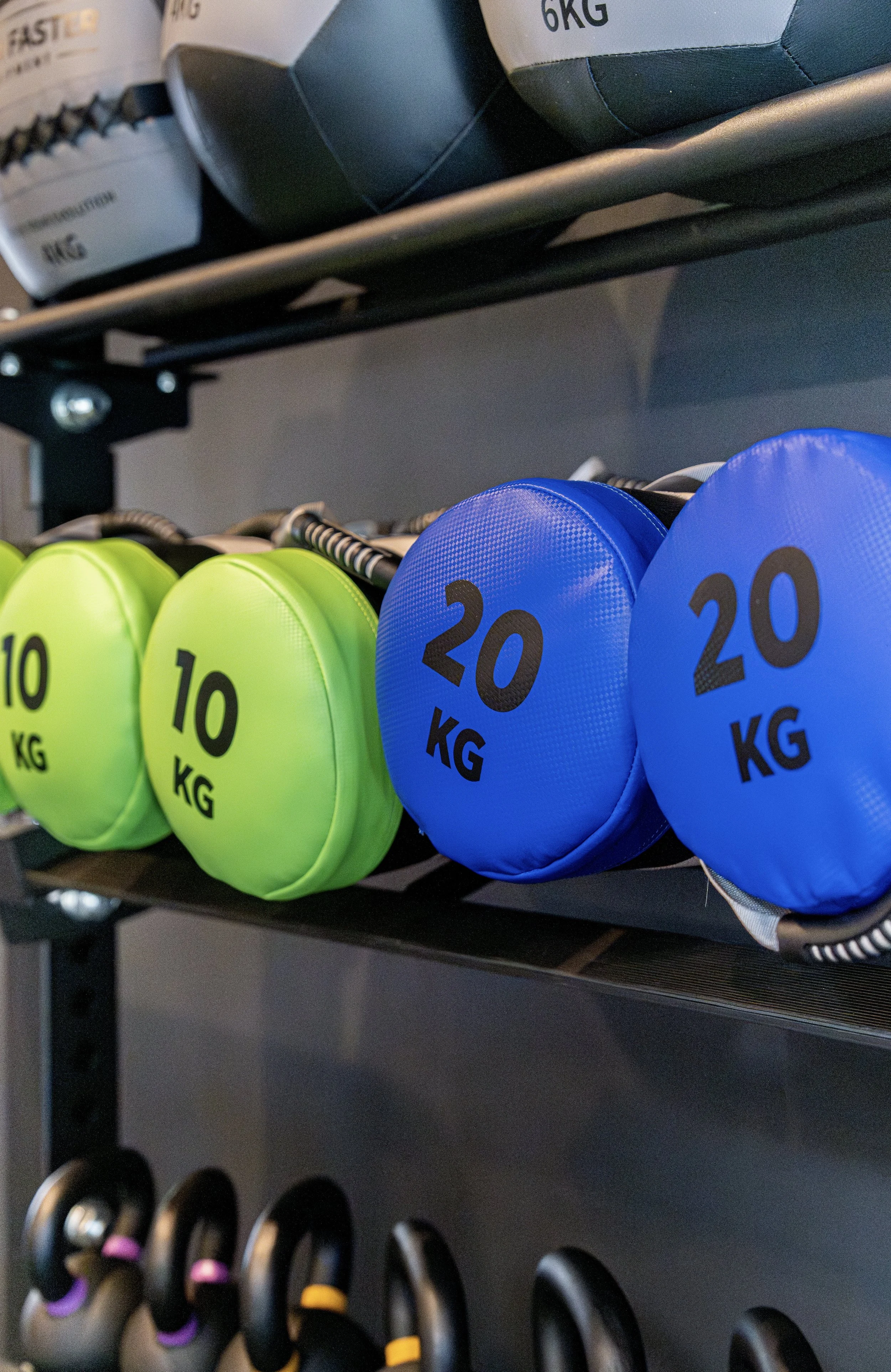 Colorful medicine balls on a metal gym rack, labeled with weights of 10 and 20 kilograms, with other fitness equipment visible below.