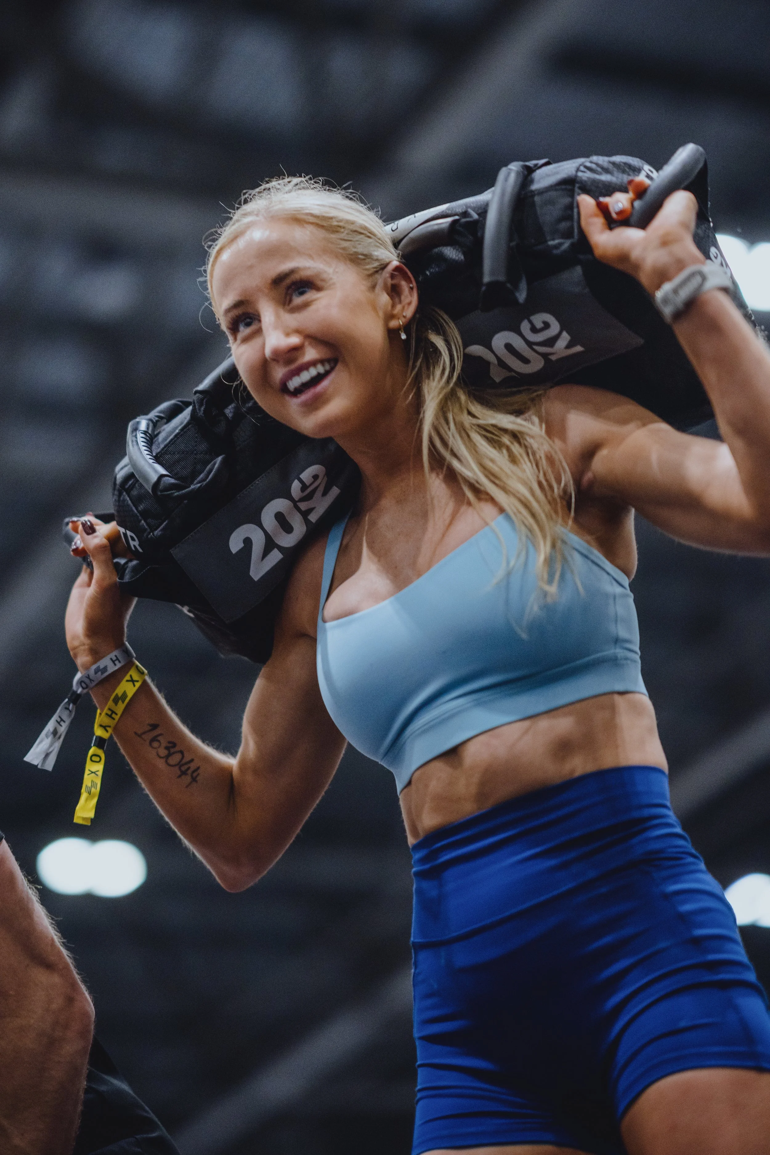 A woman in athletic clothing carries a heavy bag on her shoulders in a gym.