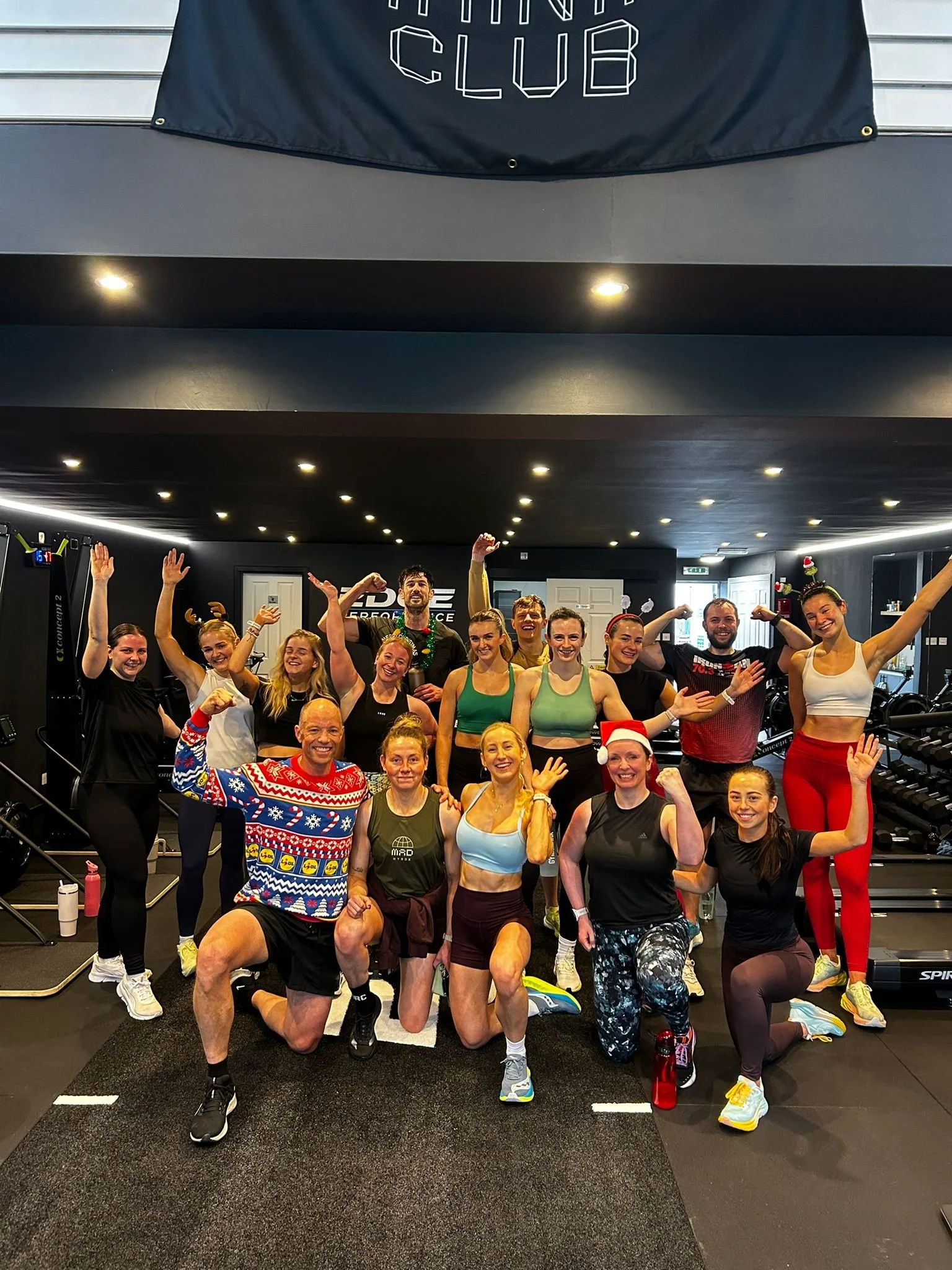 Group of people celebrating after a workout in a gym, some wearing holiday-themed clothing and accessories.